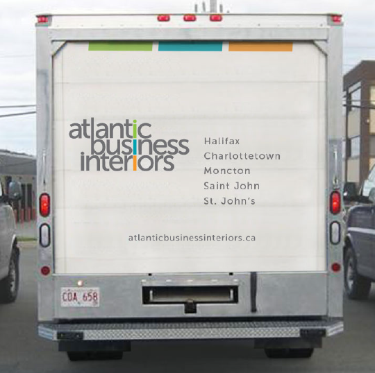 Rear view of a white delivery truck with Atlantic Business Interiors logo, listing locations Halifax, Charlottetown, Moncton, Saint John, St. John's, and website atlanticbusinessinteriors.ca.