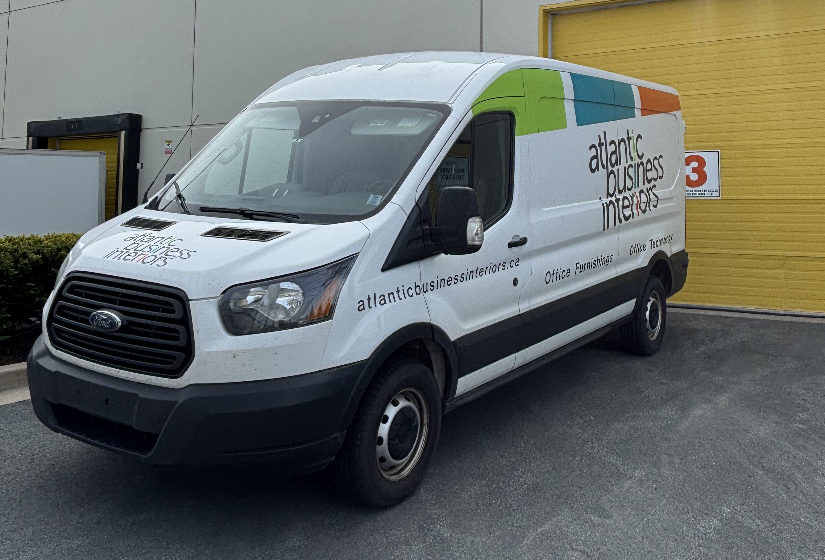 White Ford cargo van with 'atlantic business interiors' branding parked in front of a yellow industrial door.