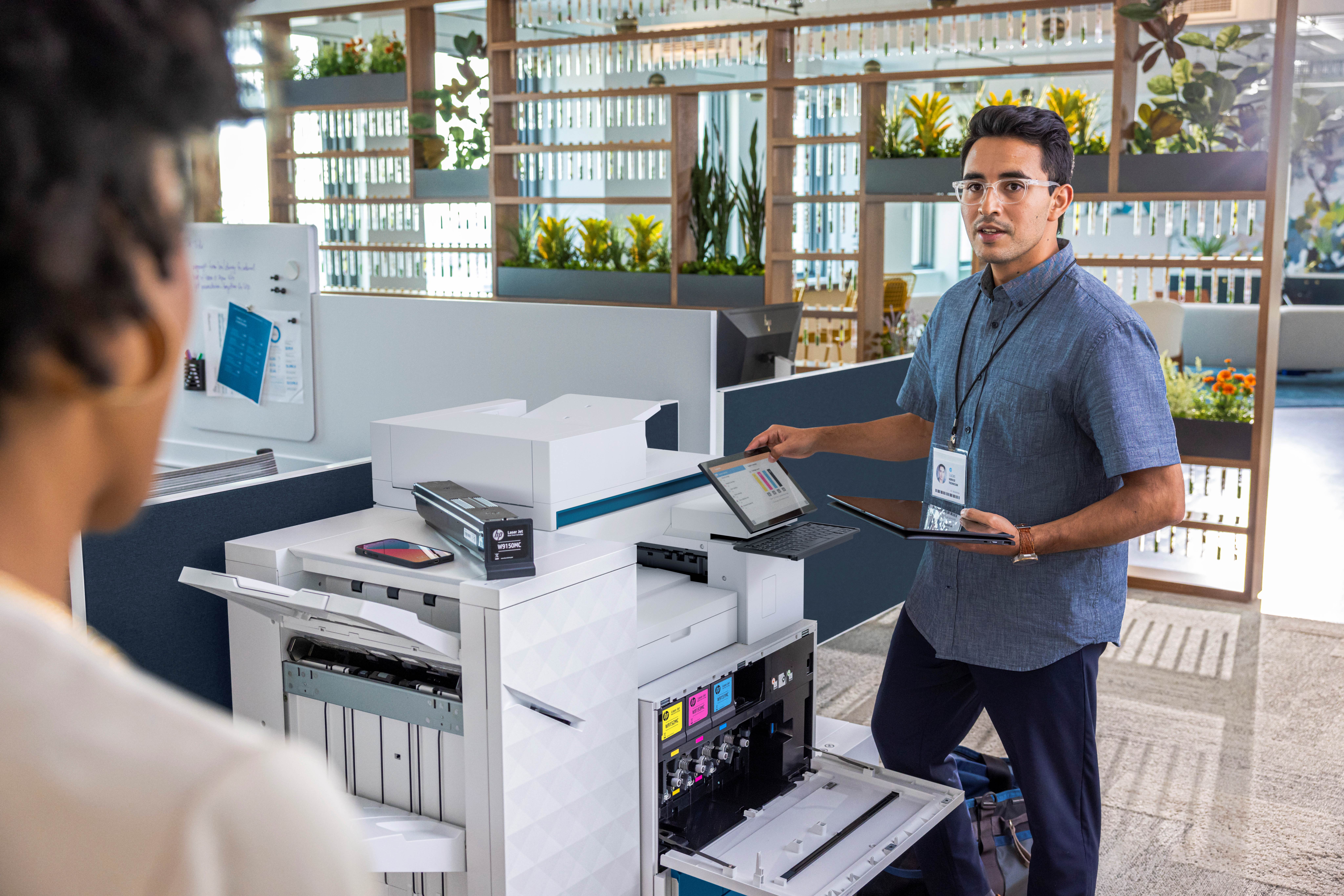 Man in blue shirt holding a tablet and standing by an open HP printer in a modern office, talking to a woman.