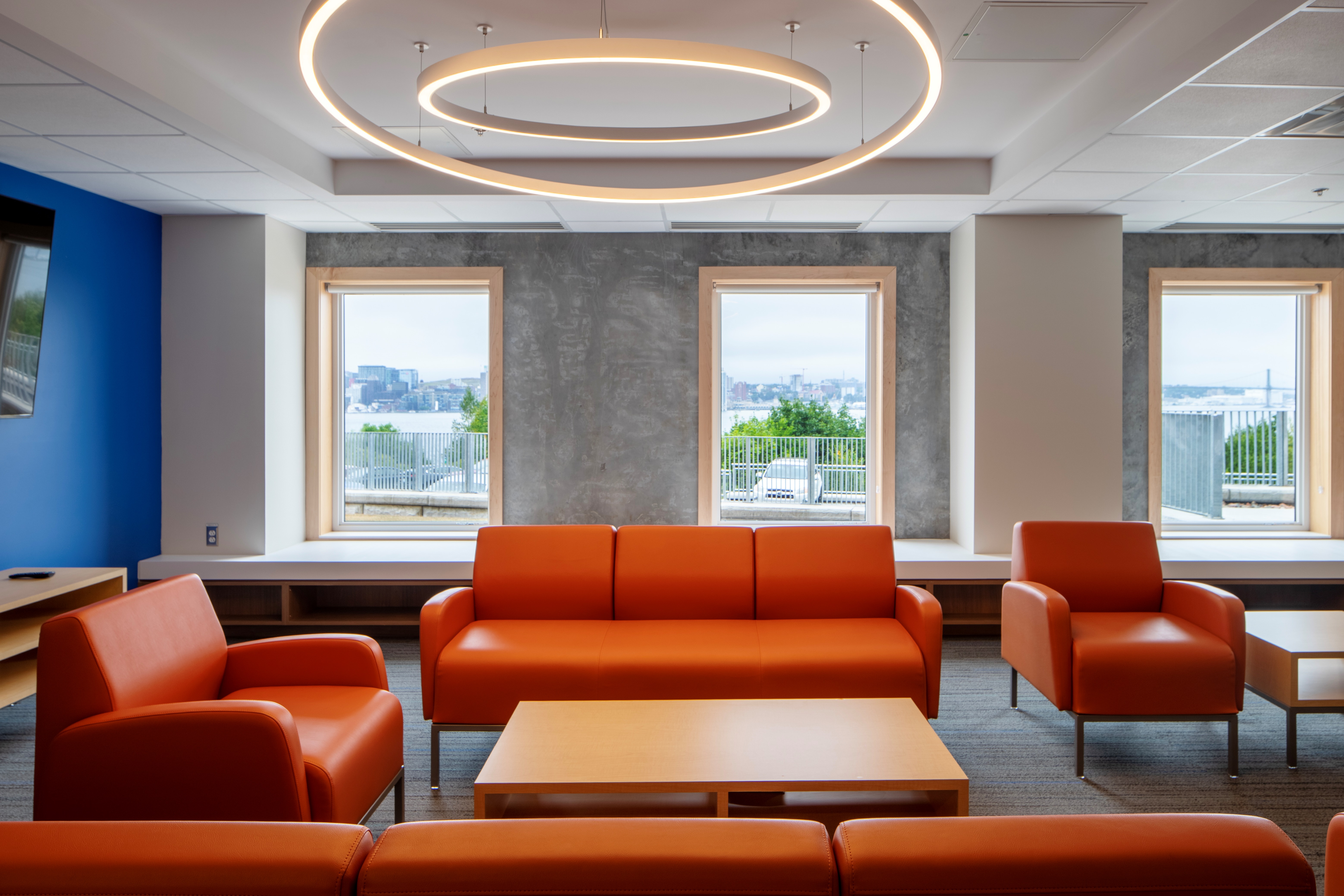 Modern office lounge with orange leather sofas, circular ceiling lights, and three large windows overlooking a cityscape.