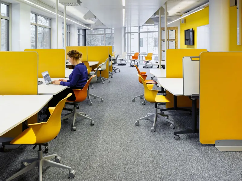 Modern office space with yellow privacy panels and chairs, featuring a woman working on a laptop.
