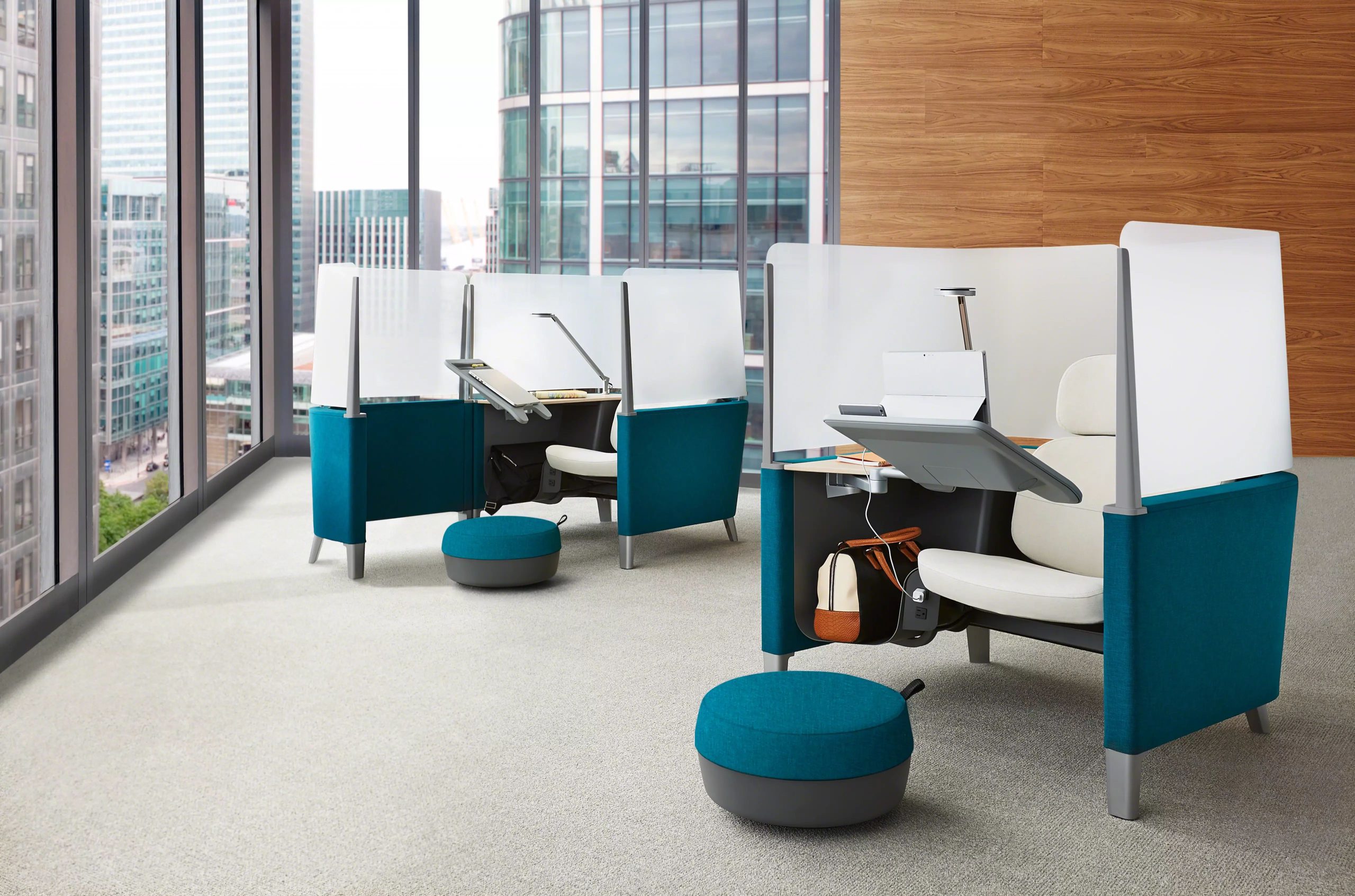 Modern office workspace pods with teal fabric panels, white cushioned chairs, adjustable desks, and footstools by large windows overlooking city buildings.