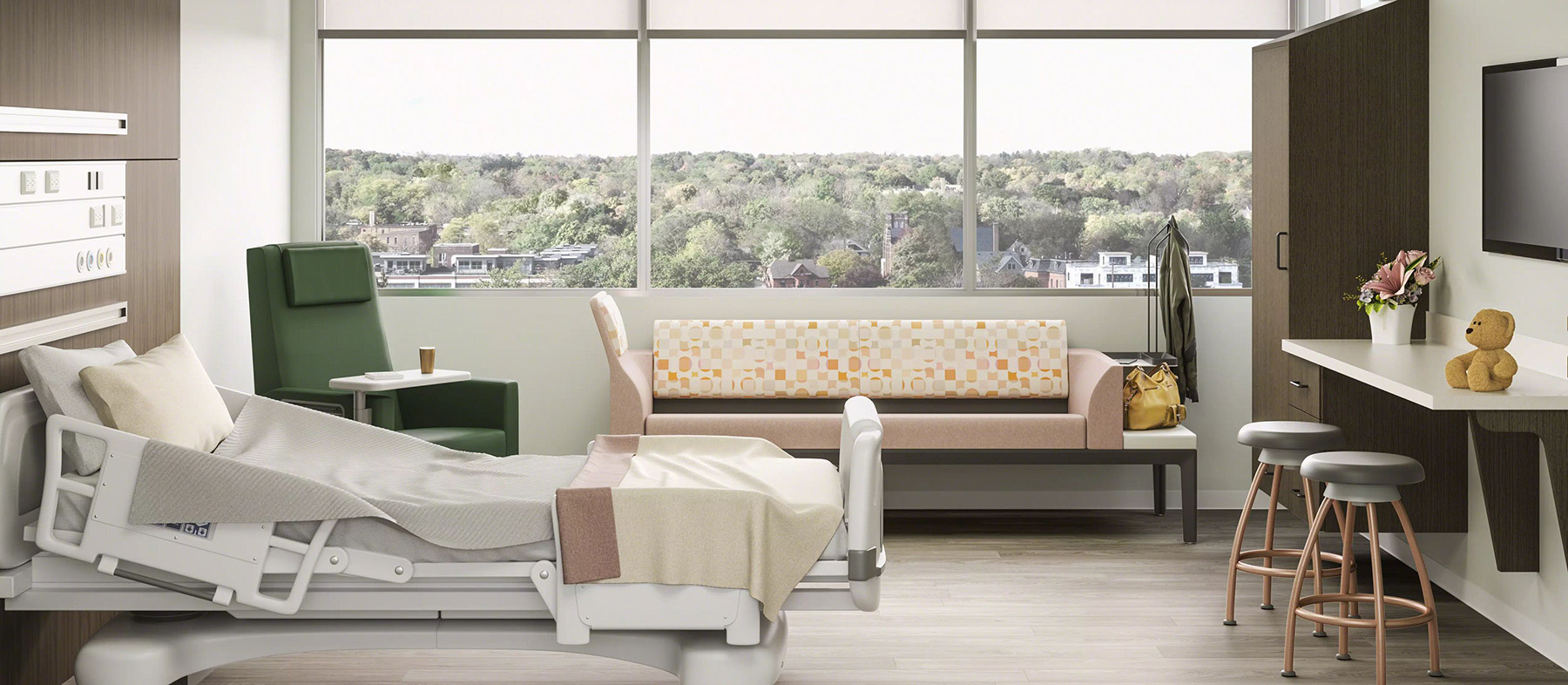 Modern hospital room with adjustable bed, green recliner, patterned sofa, coat rack, and countertop with stools, a teddy bear and flowers, window overlooking trees.