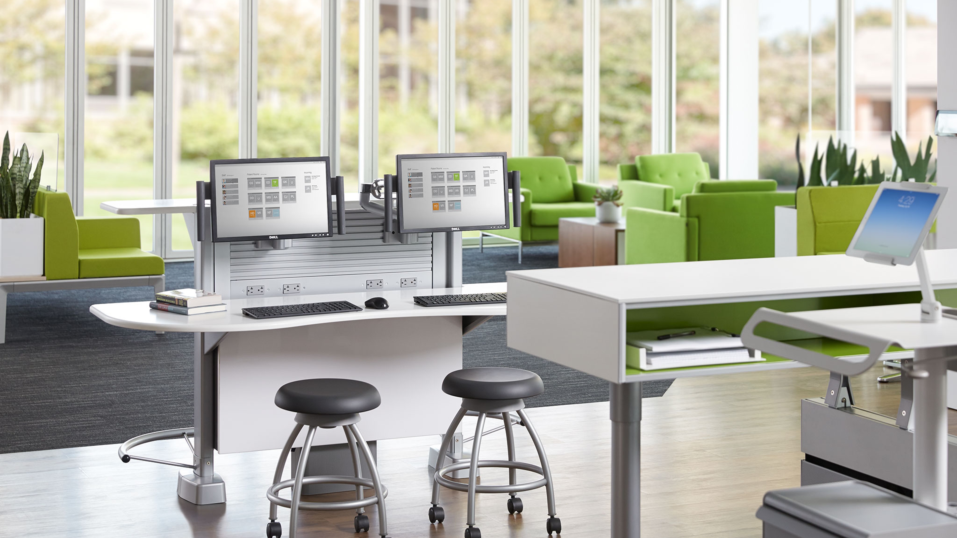 Modern office workspace with dual computer monitors, two black stools, and green lounge chairs by large windows.