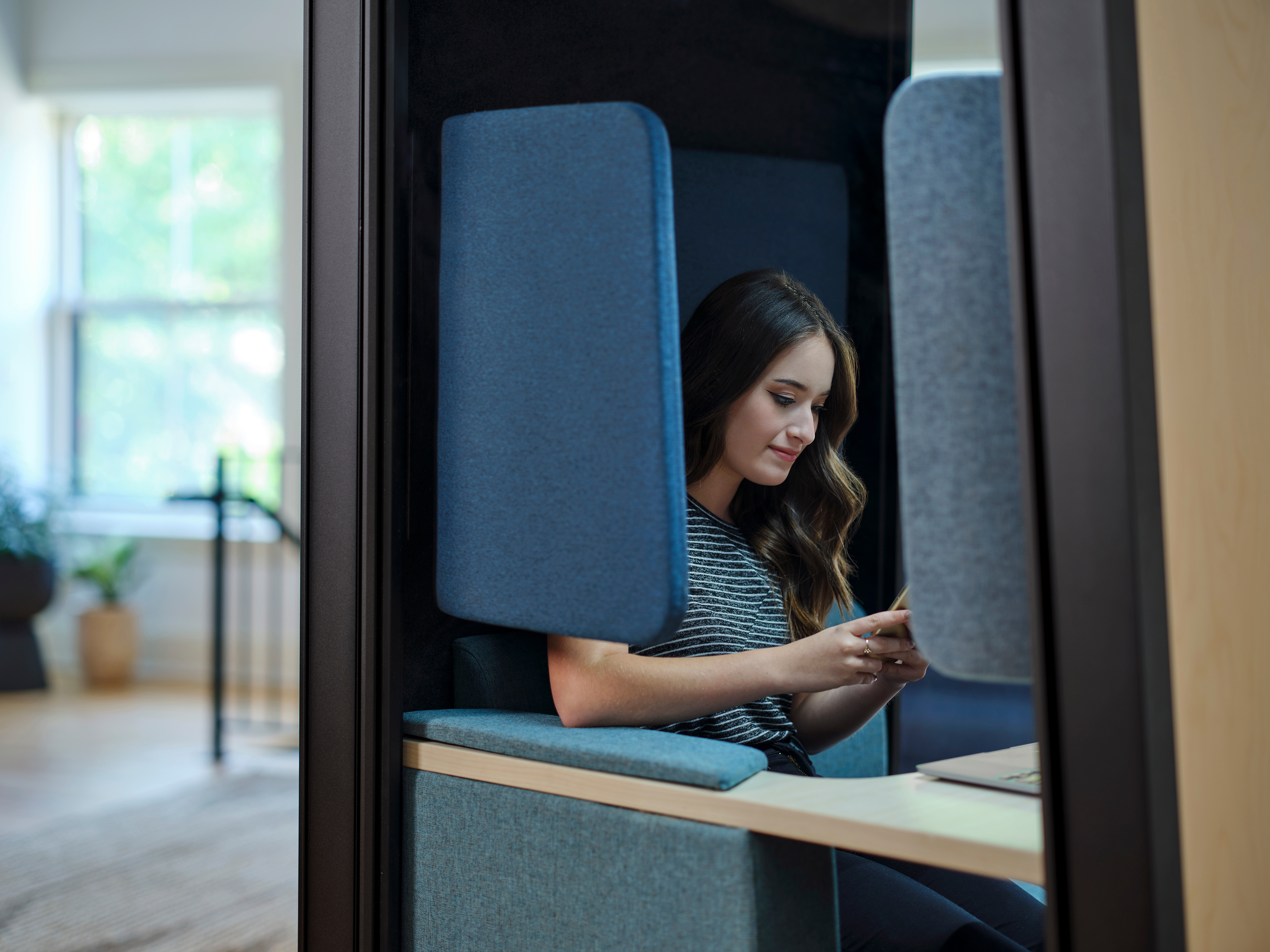 Young woman sitting in a modern blue privacy booth, looking at her smartphone with a laptop on the table beside her.