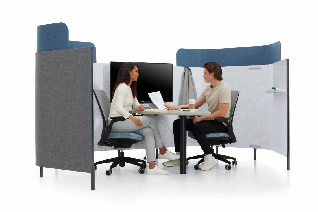 Two people sitting in a modern office pod with acoustic panels, discussing a document at a table with a monitor and coffee cup.