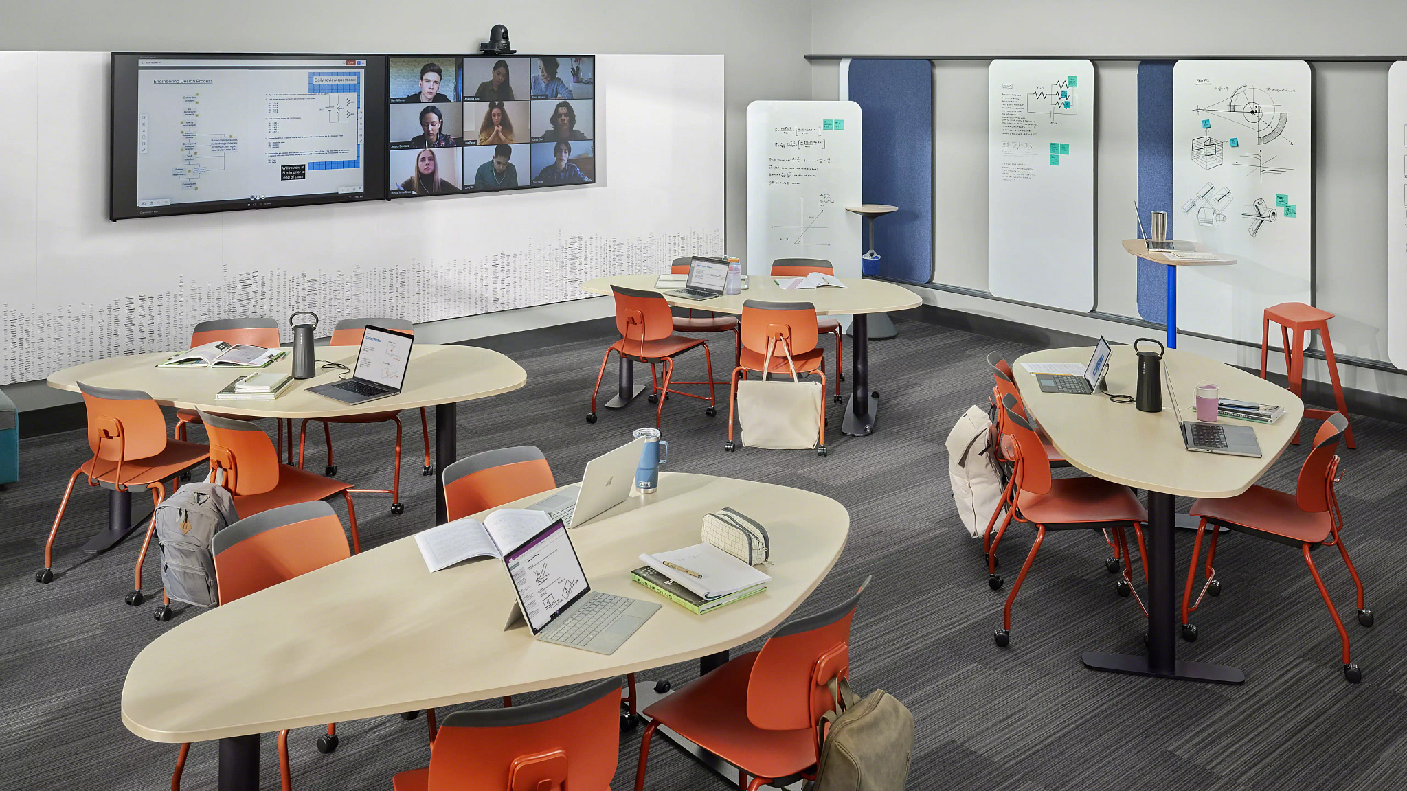Modern classroom with triple-panel video display showing engineering design and virtual meeting, tables with orange chairs, laptops, notebooks, and whiteboards with diagrams.
