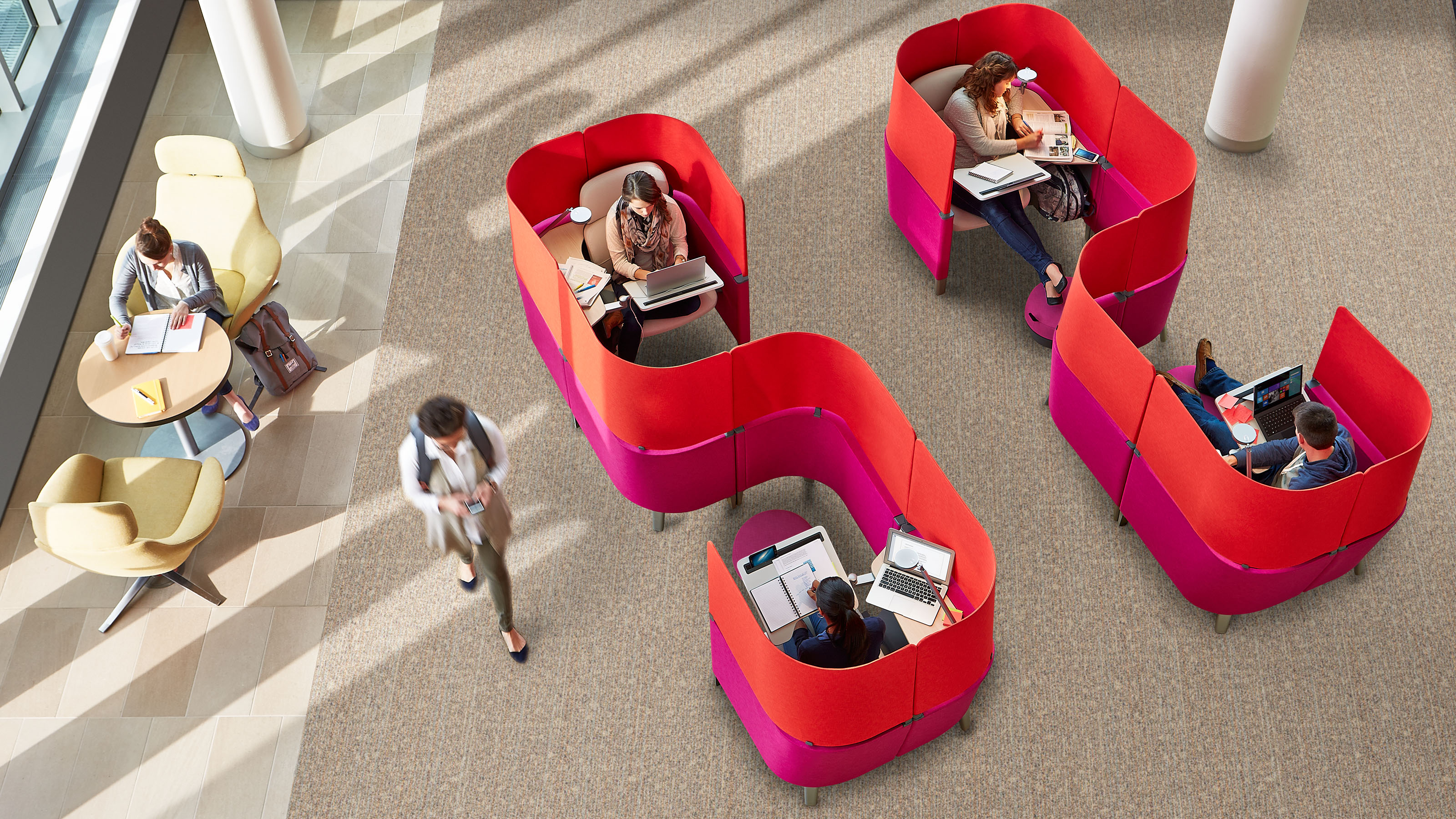 Overhead view of a modern workspace with people working on laptops and reading in privacy pods and at a round table near windows.