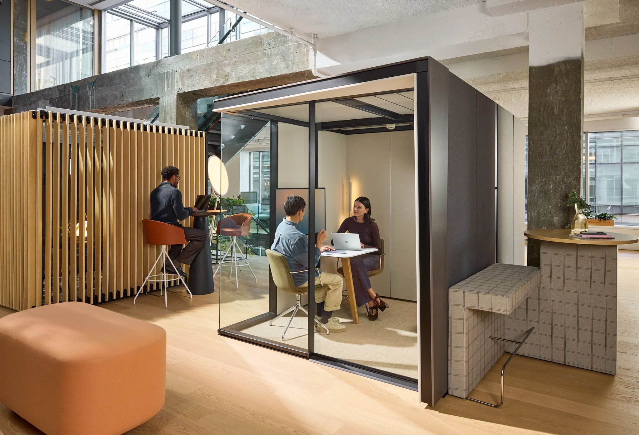 Modern office space with a glass-walled meeting pod where two people are discussing at a table, and another person working on a laptop at a standing desk nearby.