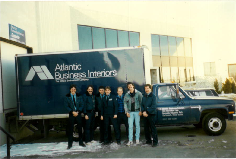 Seven men posing in front of a blue Atlantic Business Interiors truck outside a building with snow patches on the ground.