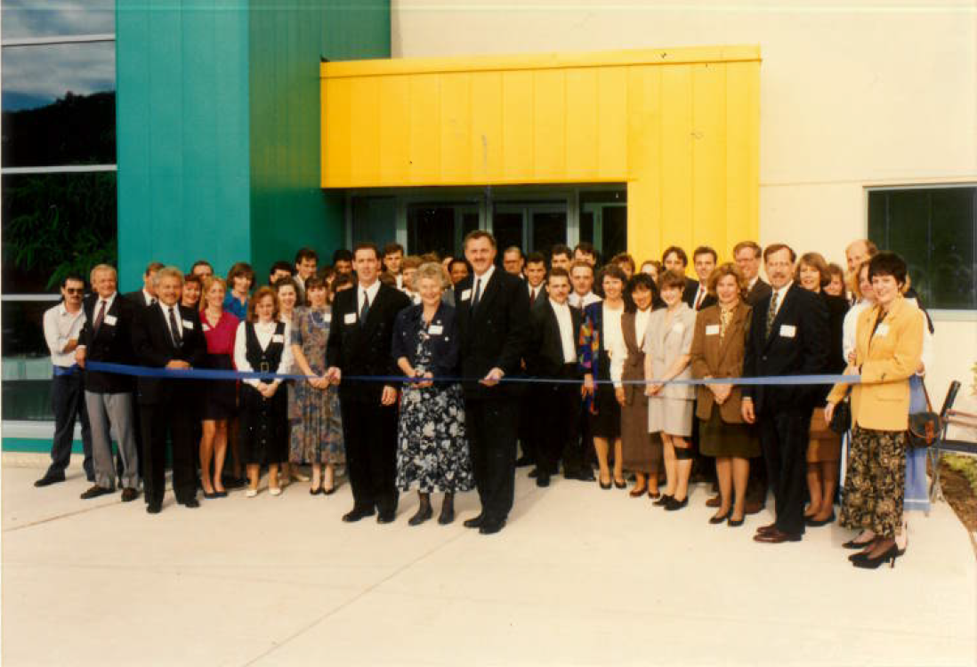 Group of professionally dressed men and women standing in front of a building with teal and yellow panels, holding a blue ribbon for a ribbon-cutting ceremony.