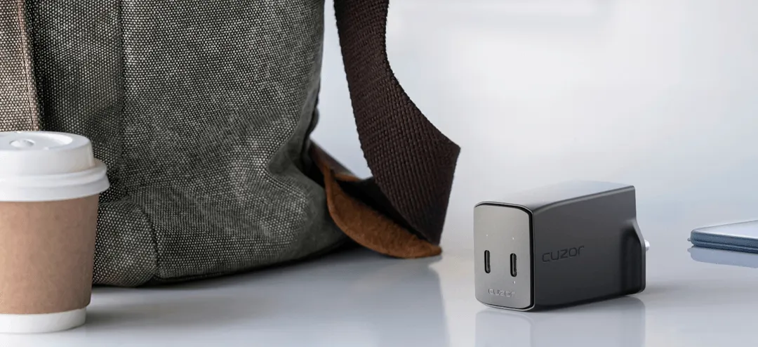 A coffee cup next to a gray backpack and a cuzor charger on a surface of the table.