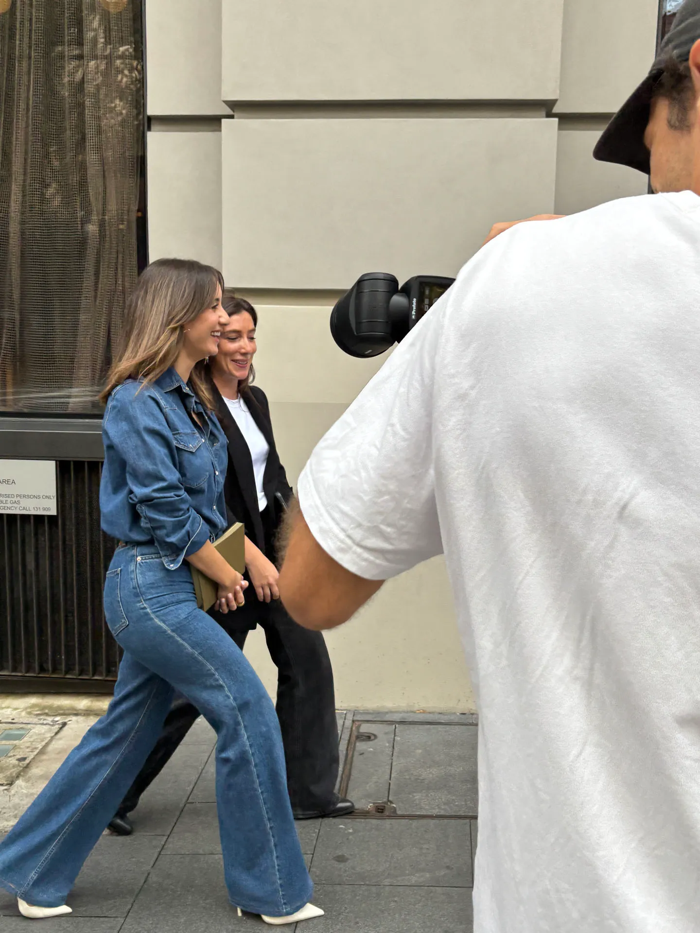 Two women walking and smiling on a city sidewalk, one in denim and holding a clutch, the other in a black blazer, while a man in a white shirt photographs them.