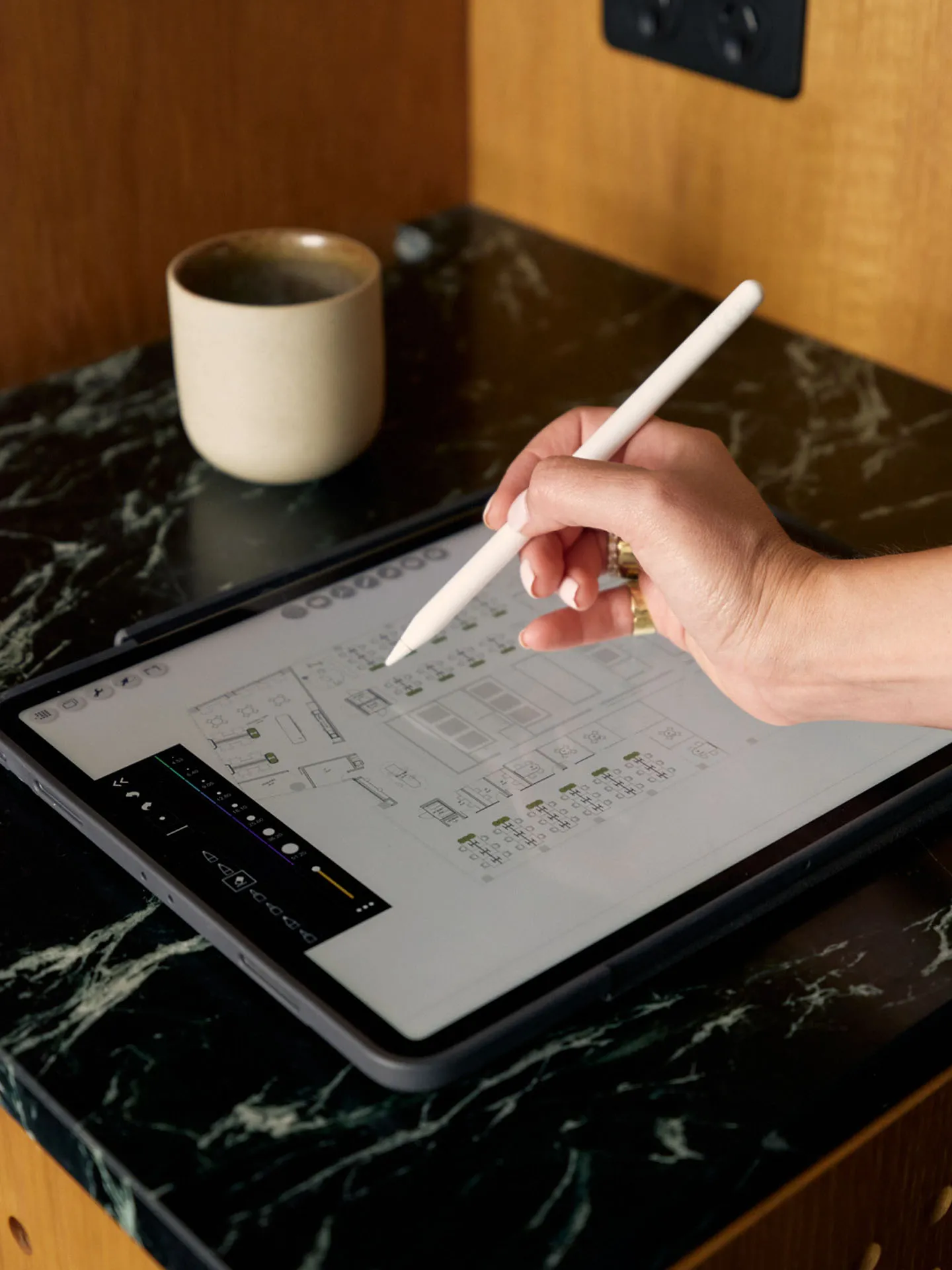 Hand holding a white stylus drawing a floor plan on a tablet next to a beige coffee cup on a black marble surface.