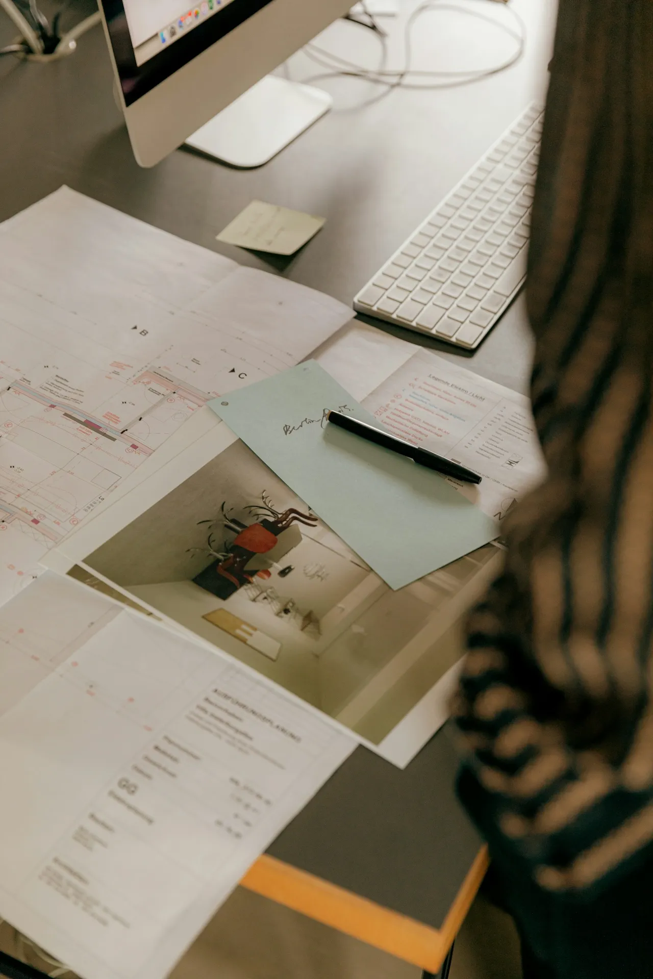 Desk with architectural plans, a photo, a pen on a blue sheet, a keyboard, and a computer monitor with visible cables.