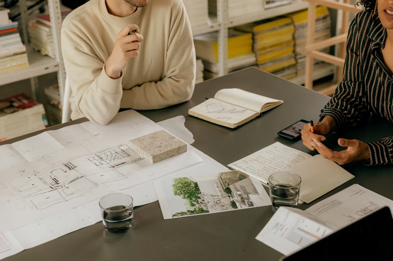 Two people at a table discussing architectural plans, sketches, and material samples with notebooks and glasses of water.