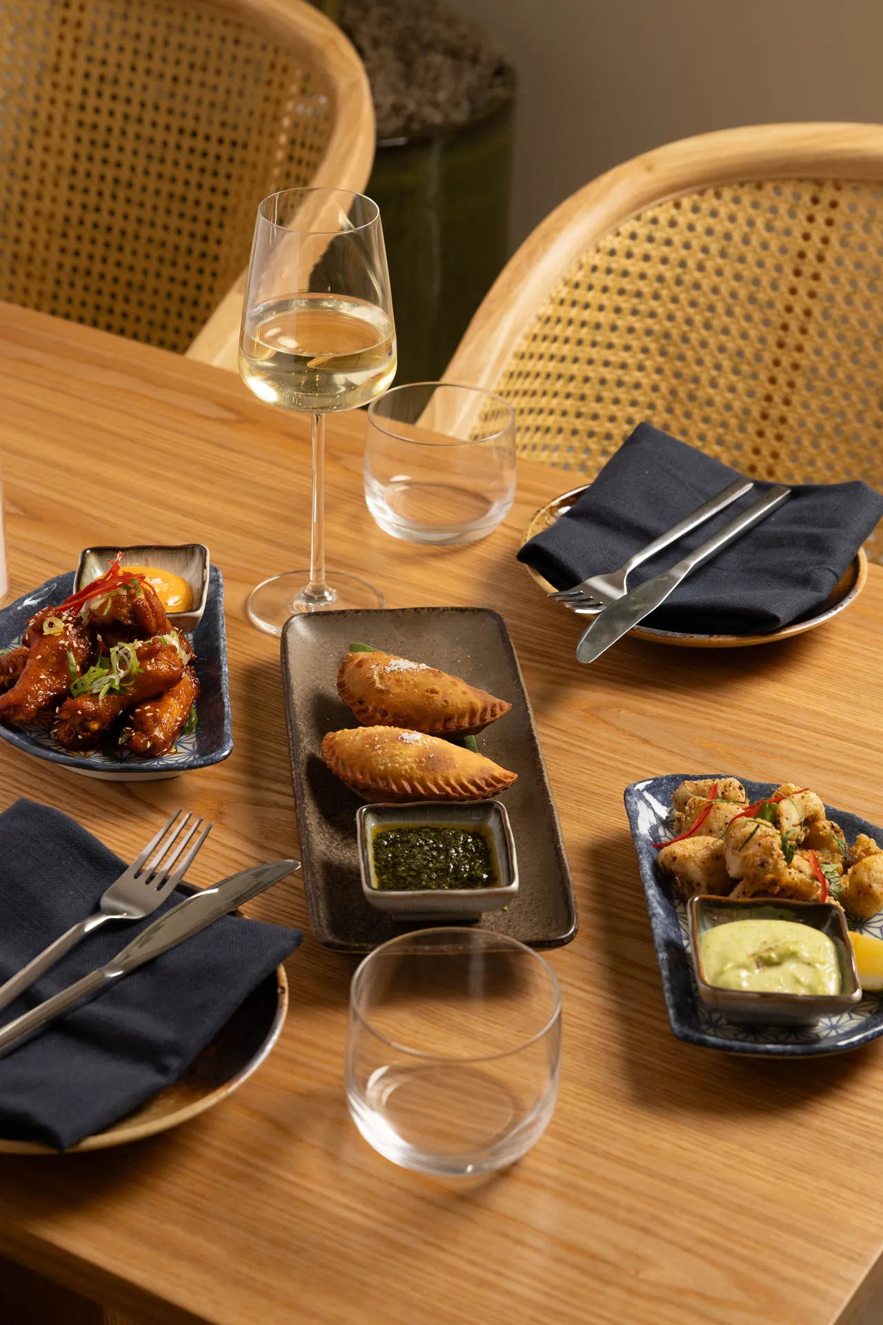 Wooden table with three plates of appetizers including chicken wings, empanadas, and fried bites with dipping sauces, a glass of white wine, and empty glasses with black napkins and cutlery.