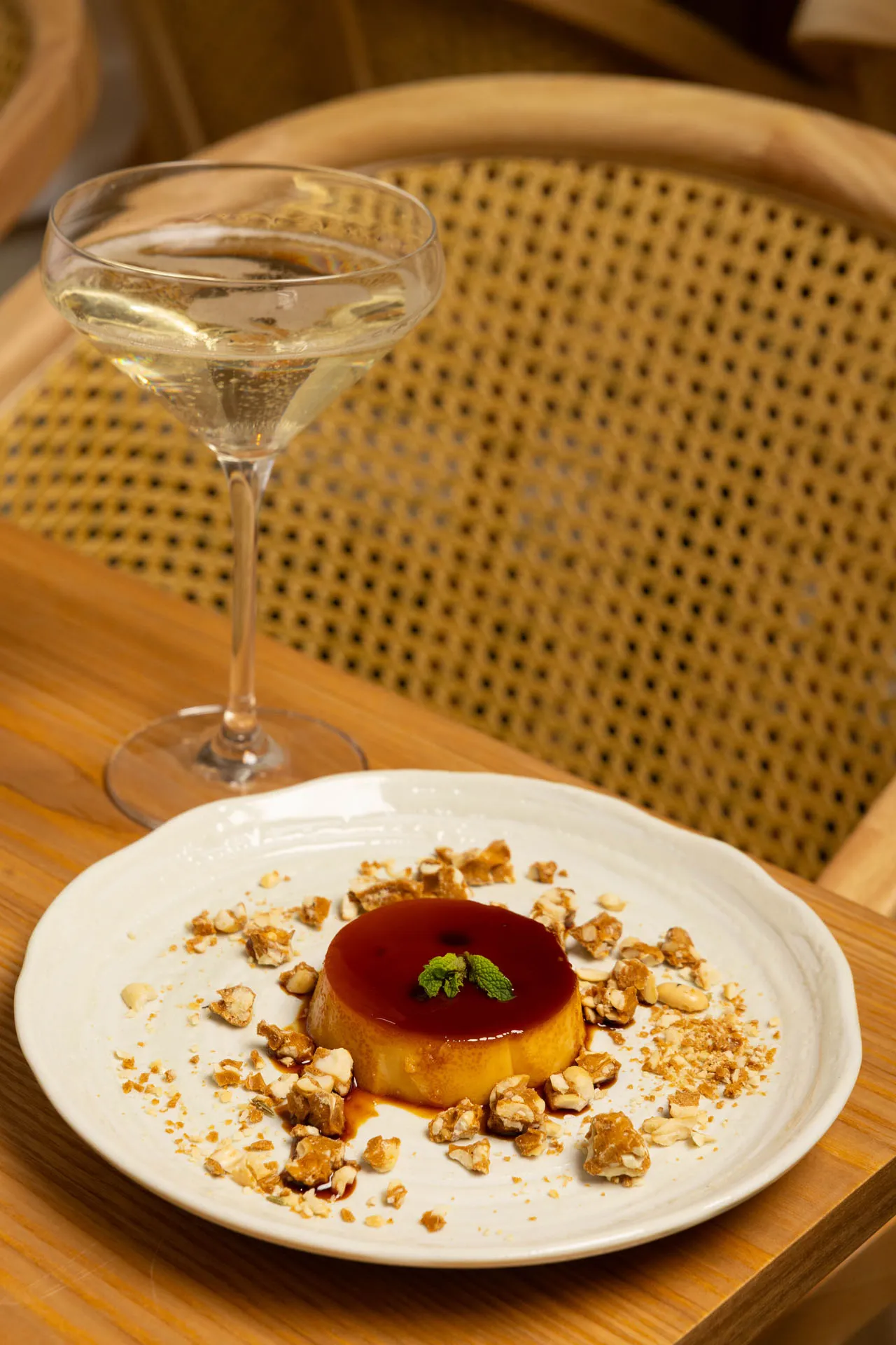 Caramel flan garnished with mint and crushed nuts on a white plate beside a glass of sparkling wine on a wooden table.