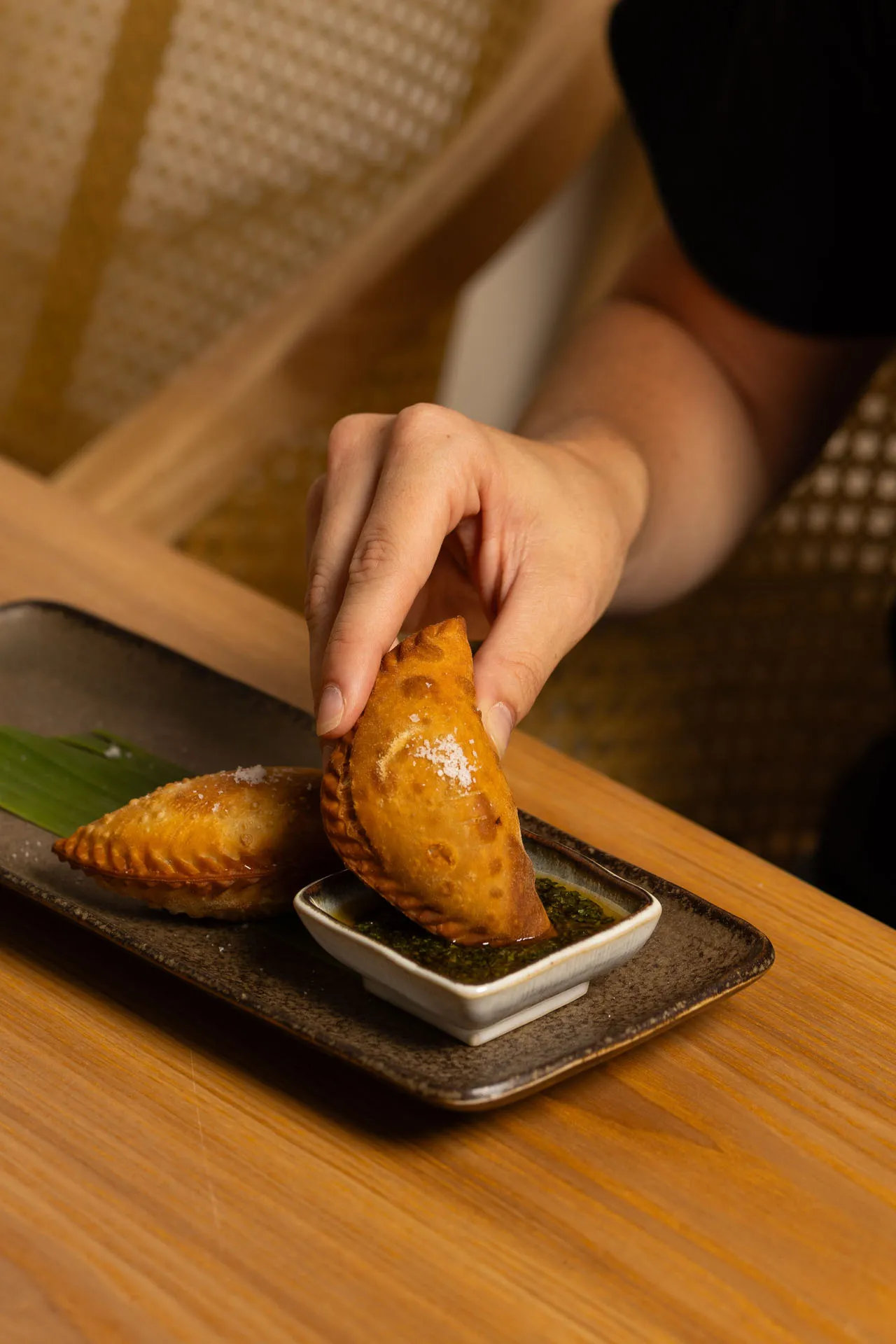 Hand dipping a golden fried empanada into a small dish of green sauce on a rectangular plate with another empanada.