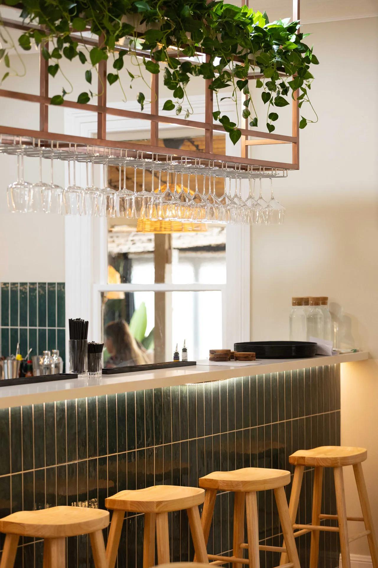 Bar area with four wooden stools, a tiled counter front, hanging wine glasses, and green plants overhead.
