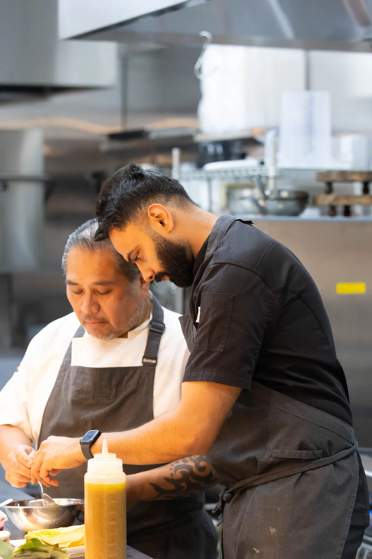 Two chefs in aprons working together in a professional kitchen, focused on preparing food in a metal bowl.