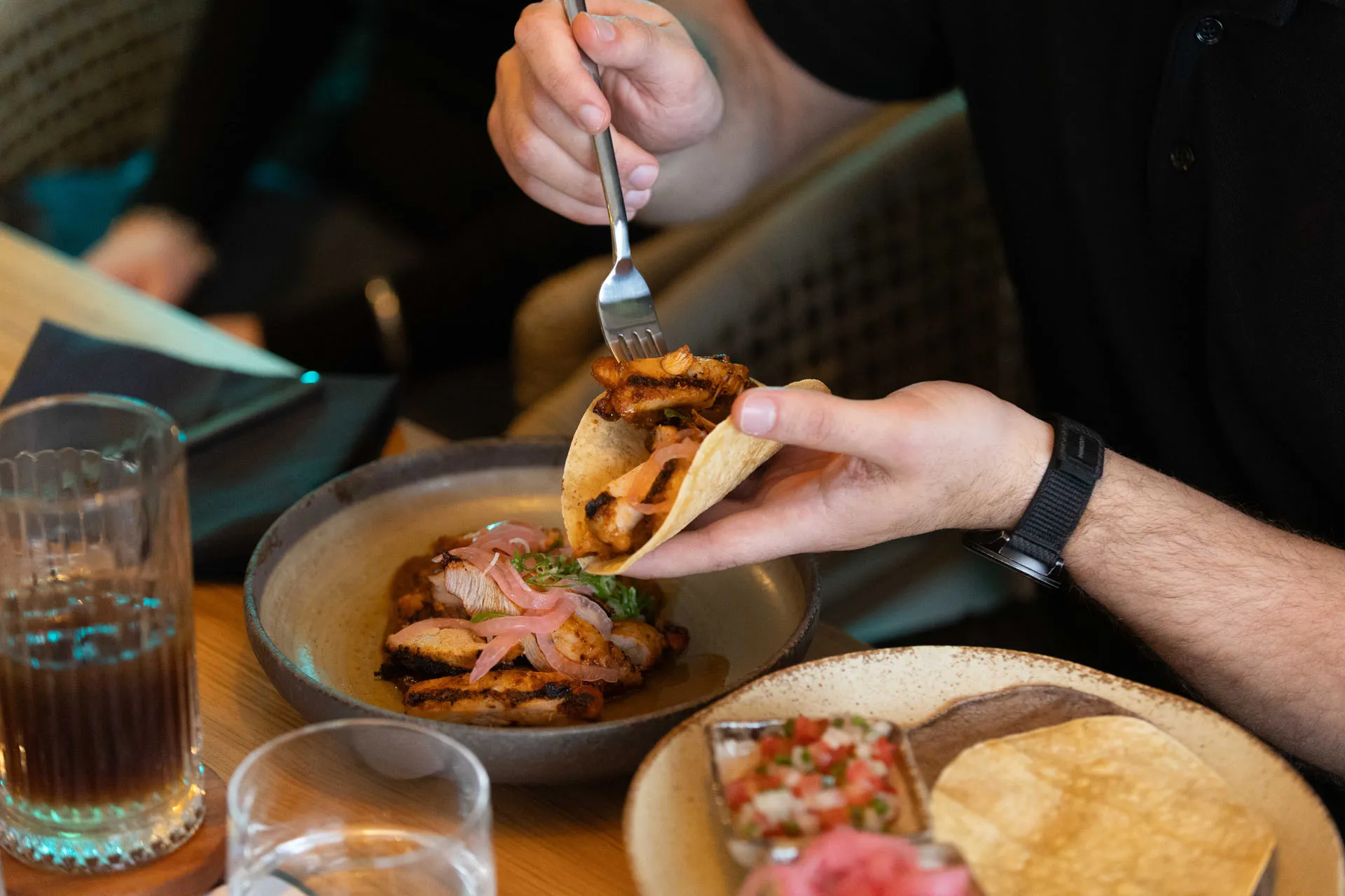 Person holding a taco filled with grilled chicken and pickled onions, with a bowl of grilled chicken and a plate of tortillas and salsa on the table.