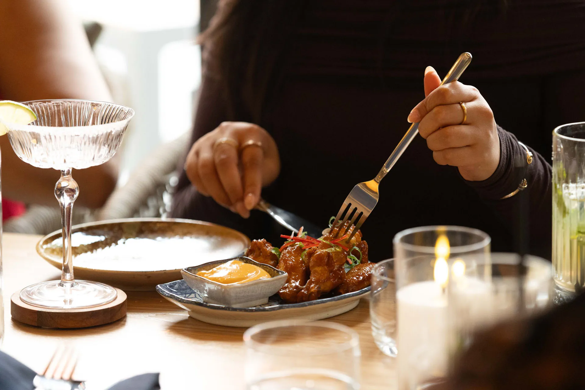 Person using fork and knife to eat glazed chicken wings with dipping sauce on a blue plate at a restaurant table.