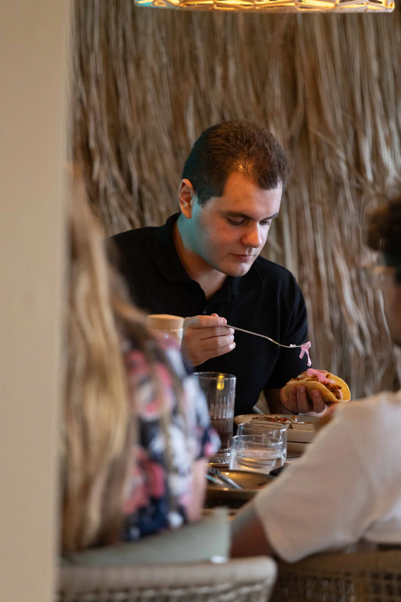 Man in black shirt adding pink pickled onions to a sandwich while seated at a table with drinks and plates.