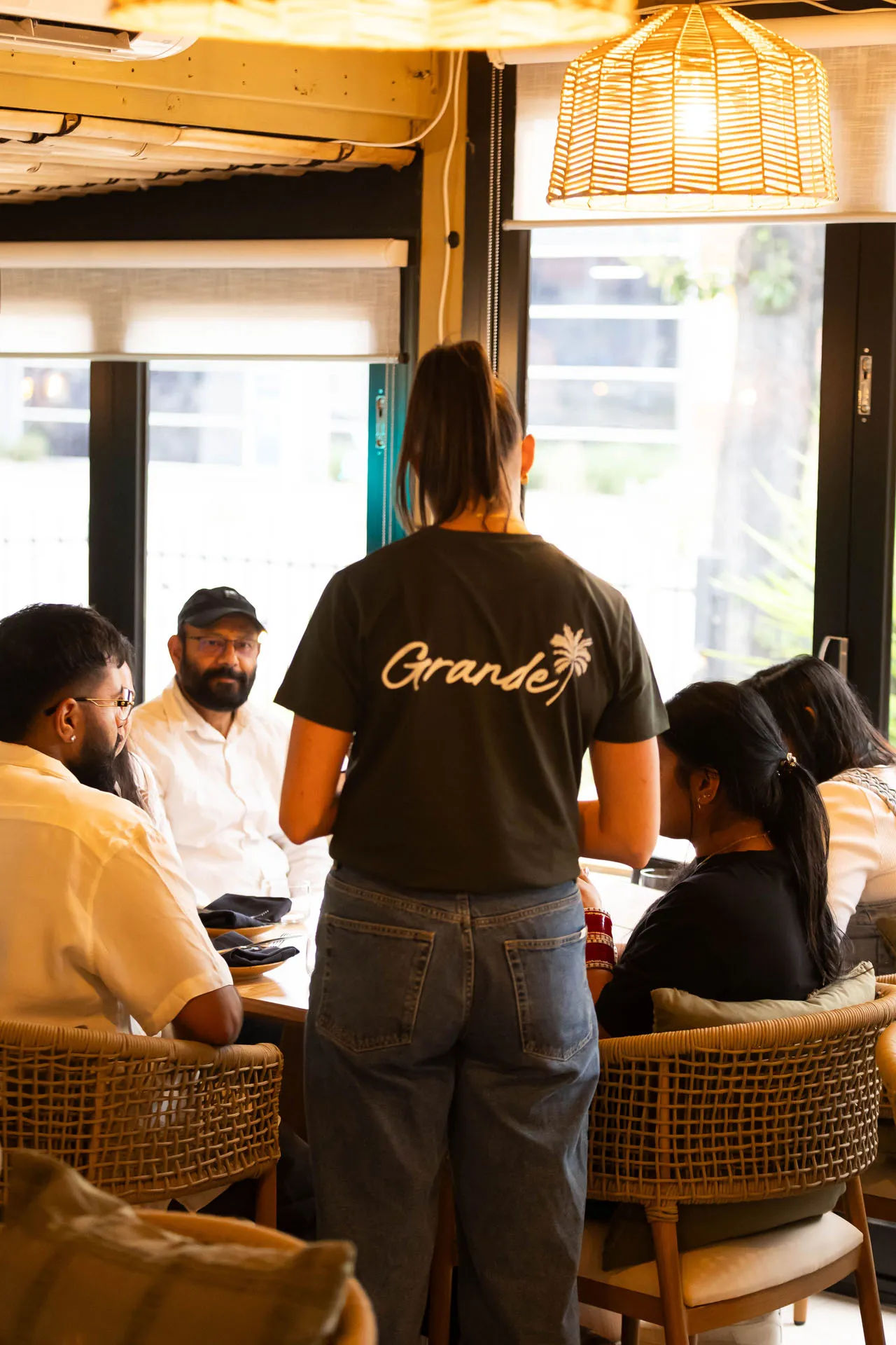 Waitress wearing a 'Grande' t-shirt serving a group of people seated at a restaurant table near large windows.