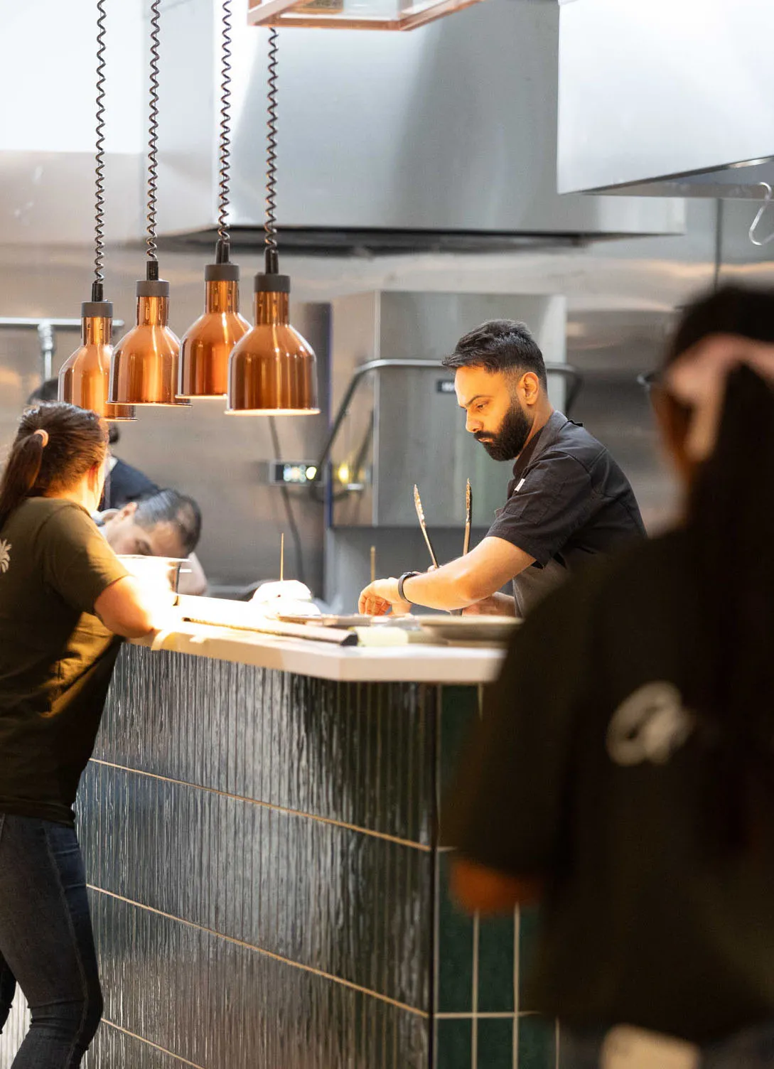 Man with a beard working behind a kitchen counter with hanging copper lights while a woman leans on the counter talking to him.