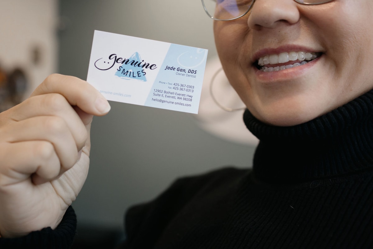 Person wearing glasses displaying a Genuine Smiles business card while smiling at the camera.