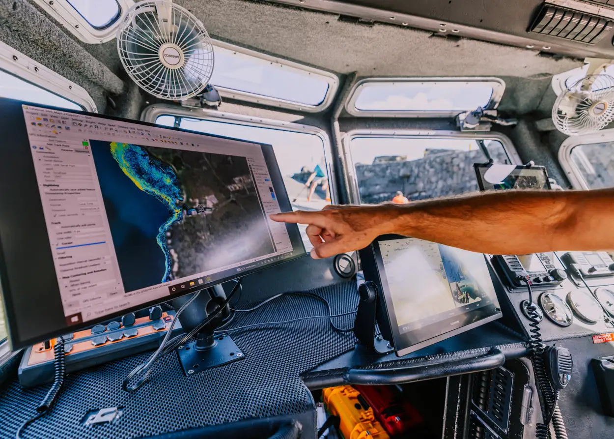 Person pointing at a coastal map displayed on a computer screen inside a boat cockpit.