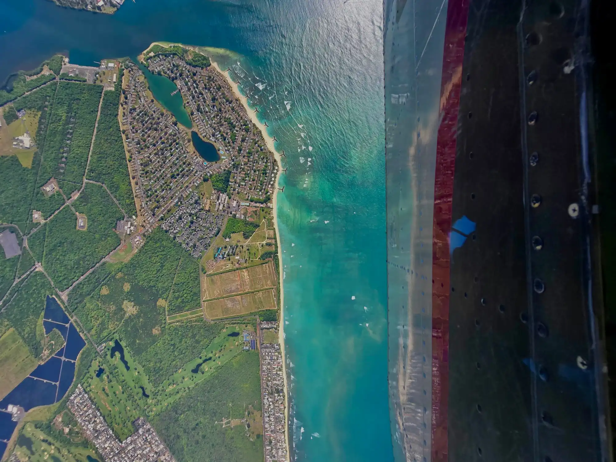 Aerial view of a coastline with blue ocean, sandy beach, residential areas, and greenery from airplane window.