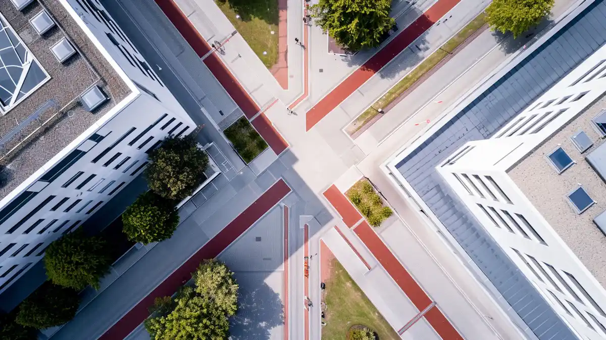 Aerial view of an intersection with pedestrian paths, red bike lanes, green trees, and white buildings casting shadows.