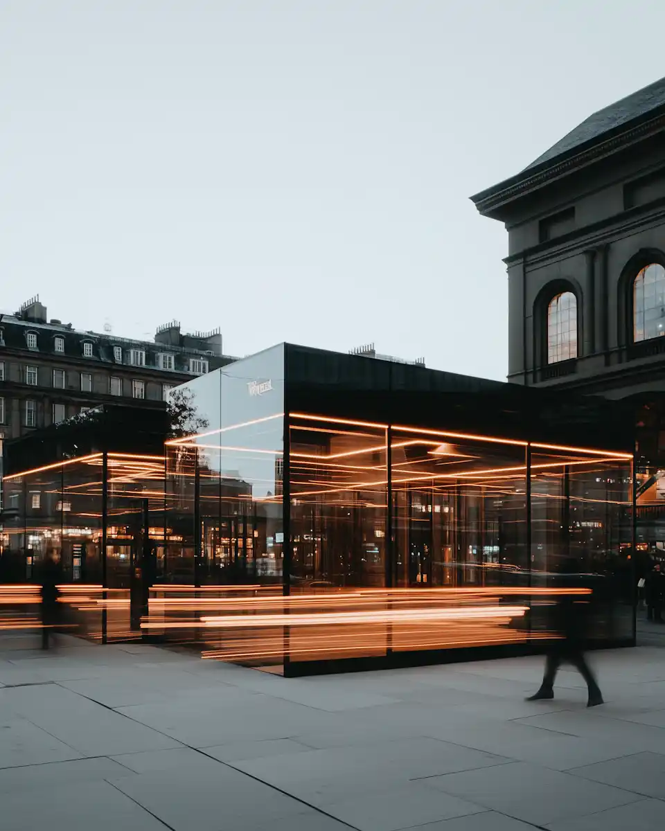 Modern glass structure with orange neon lighting lines in an urban plaza during dusk.