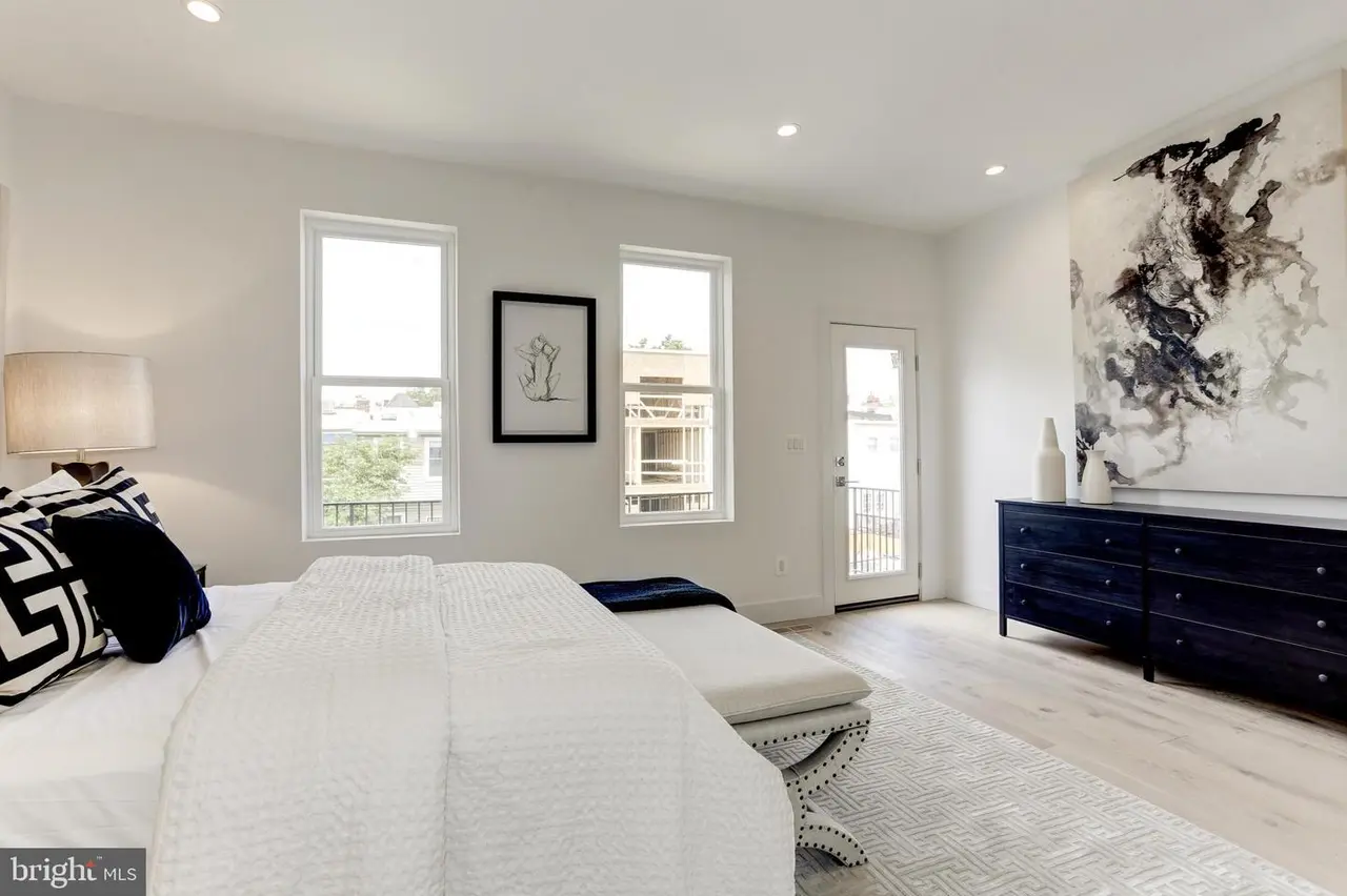Modern bedroom with white walls, two tall windows, white bedding, a bench at the foot of the bed, and a black dresser with abstract art above it.