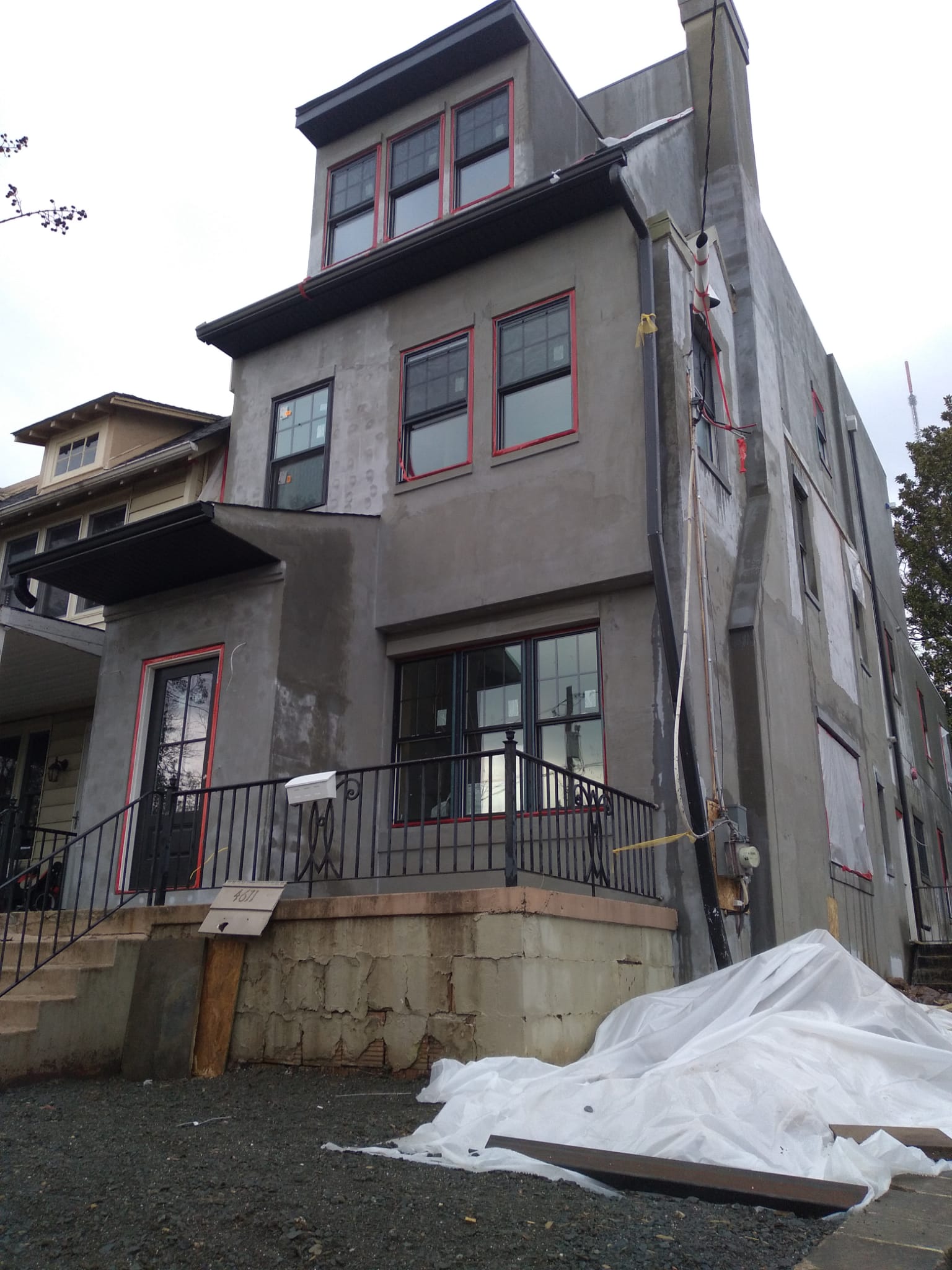 Three-story house under construction with gray exterior, red-trimmed windows, a black iron railing, and a pile of white construction tarp in front.
