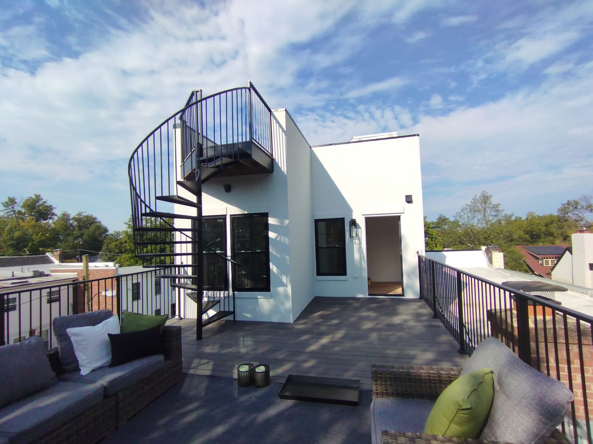Rooftop patio with gray cushioned wicker furniture, green and white pillows, black spiral staircase, and white building under a blue sky with clouds.