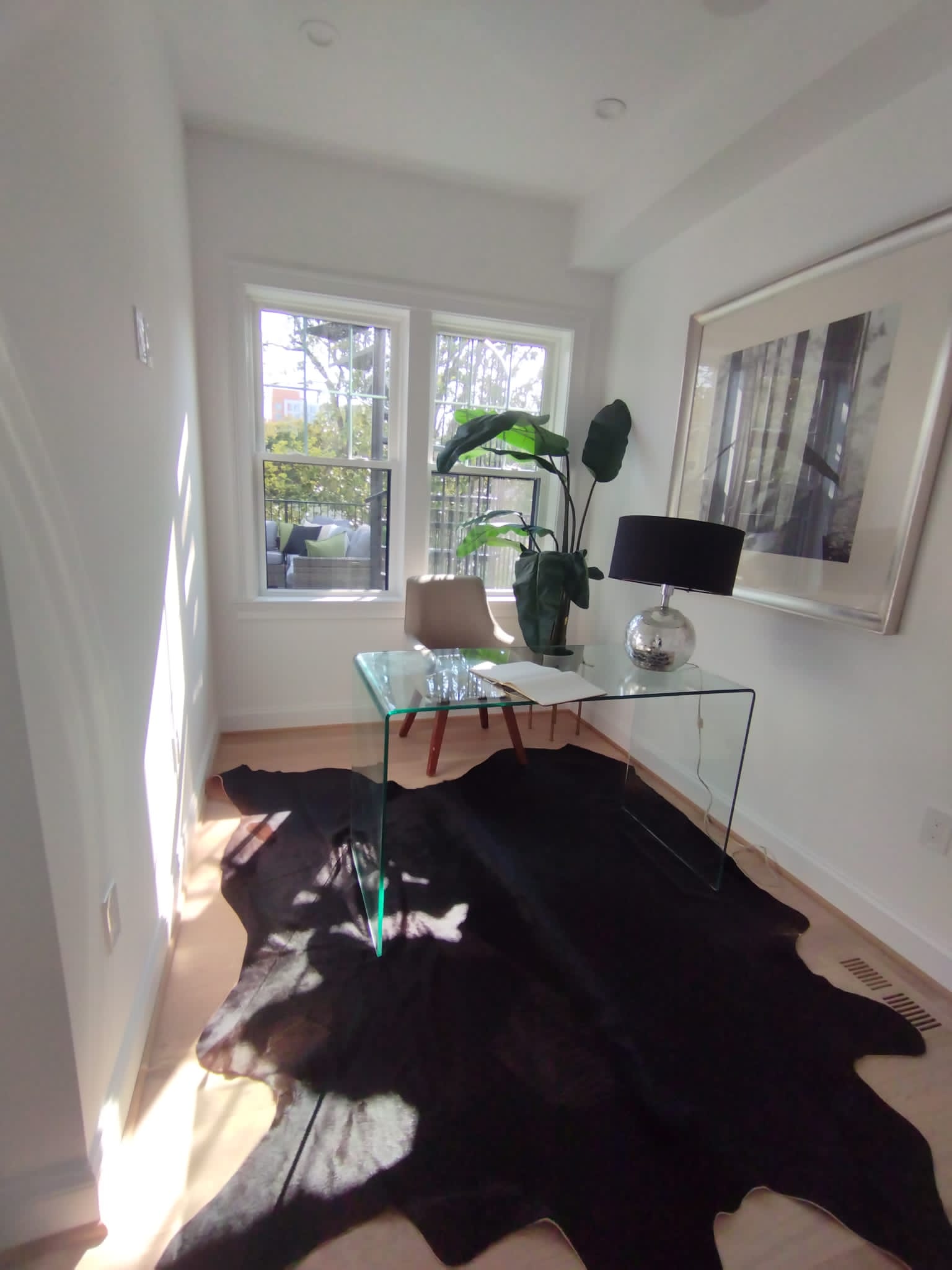 Bright minimalist home office with glass desk, beige chair, large leafy plant, black lamp, and framed forest artwork on white walls.