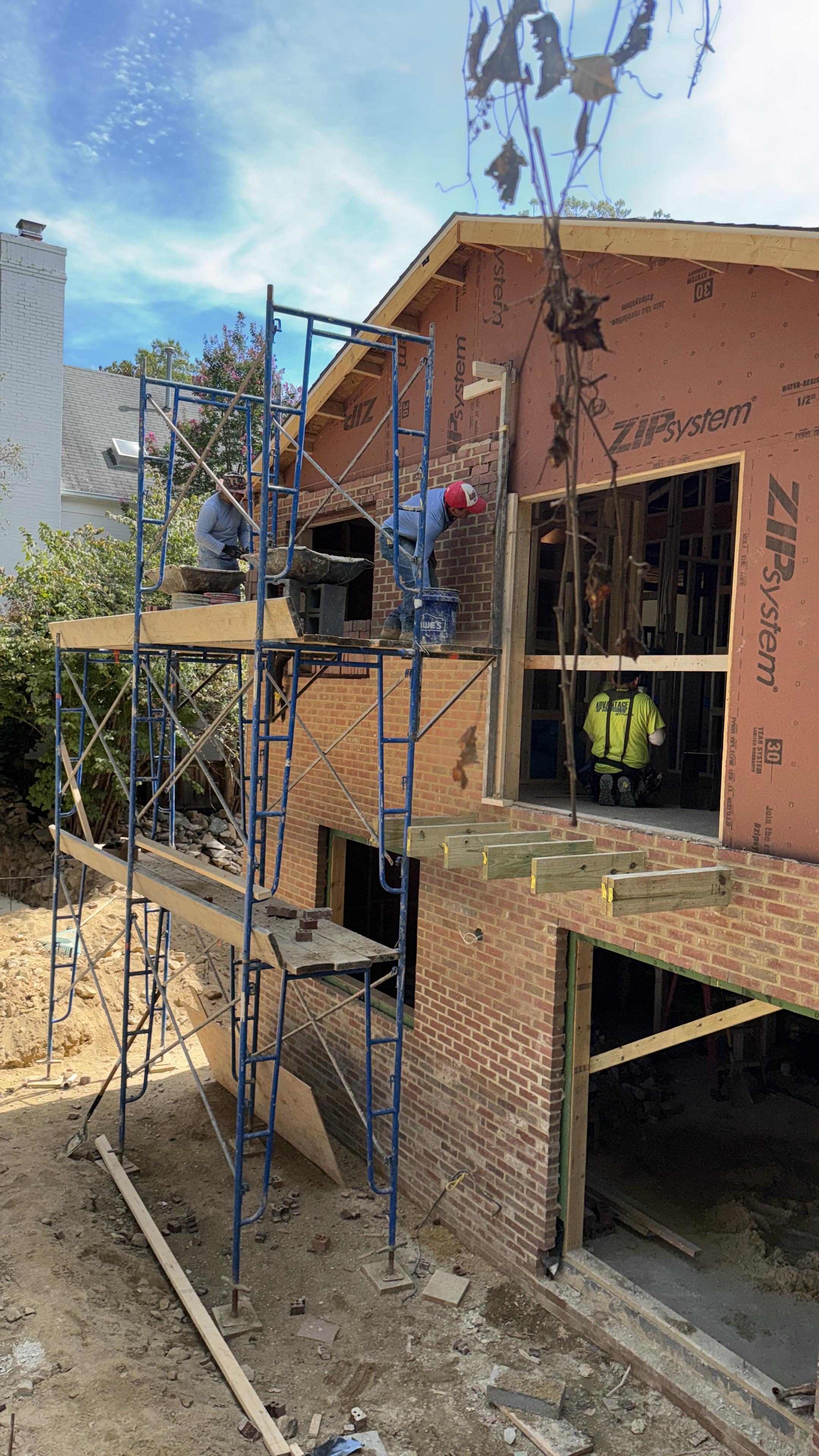 Construction workers on scaffolding building a brick exterior wall of a house under renovation.