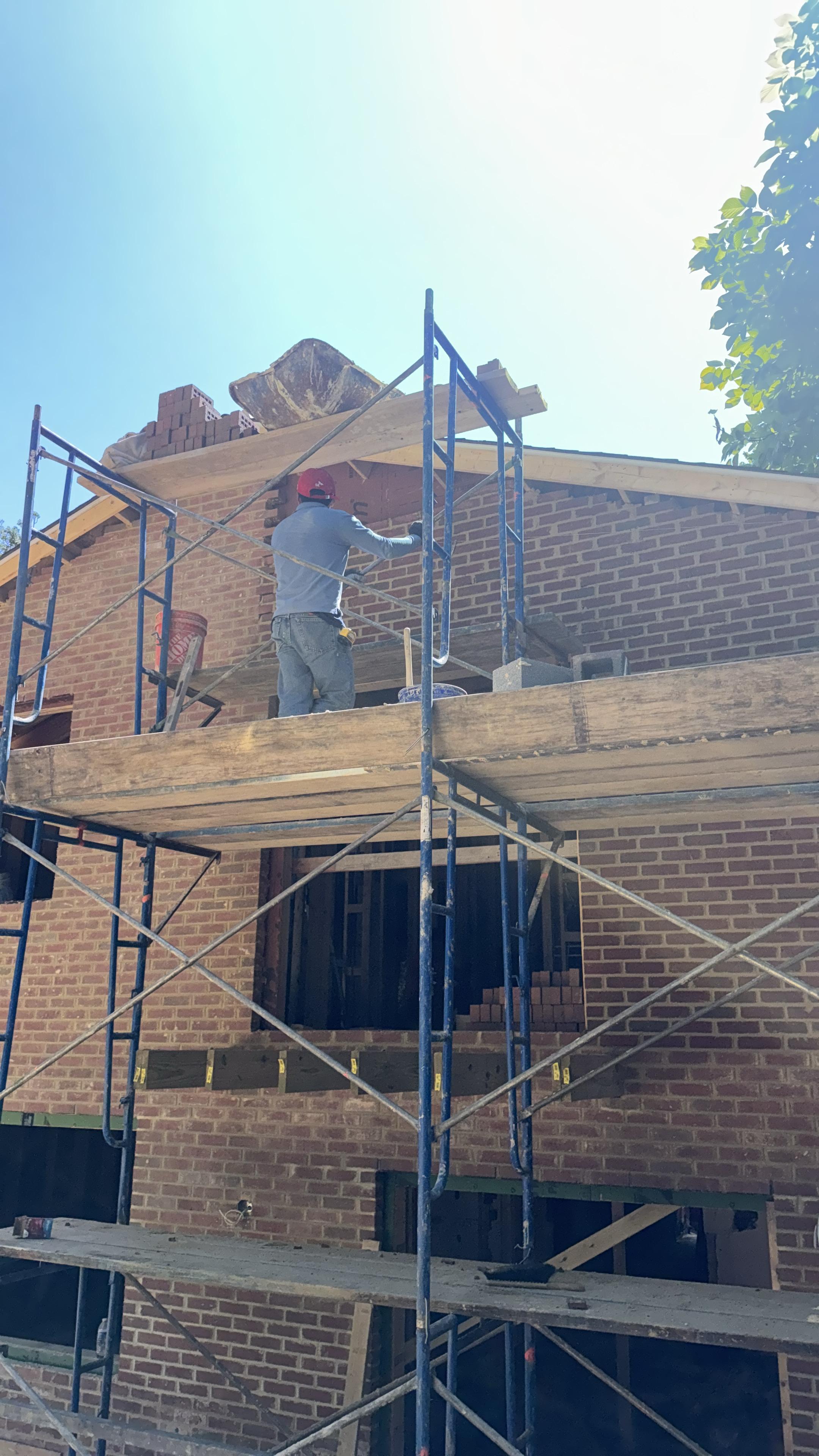 Construction worker on scaffolding building a brick wall under a bright sky.