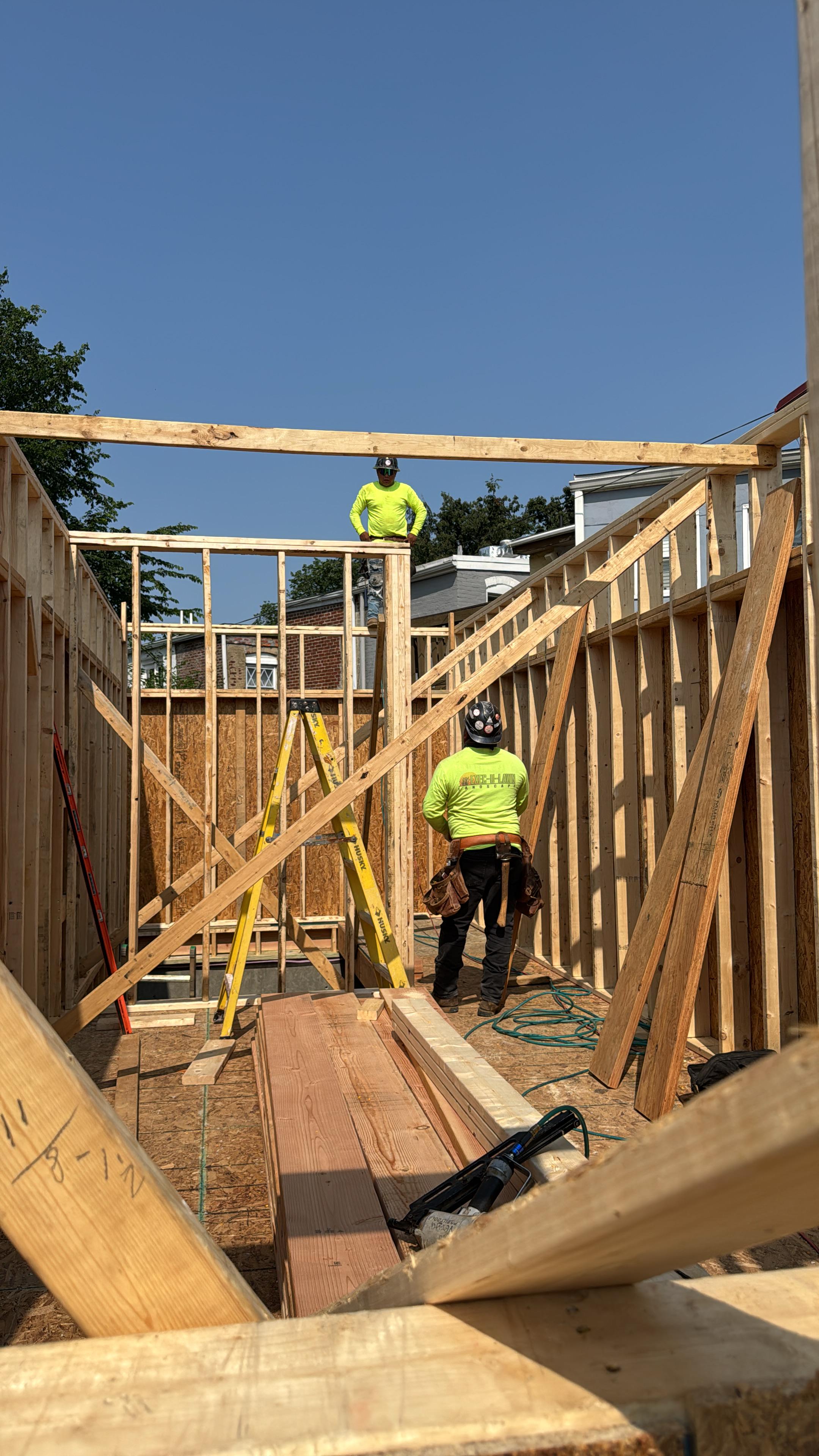 Two construction workers in neon yellow shirts building wooden framework inside a structure under a clear blue sky.