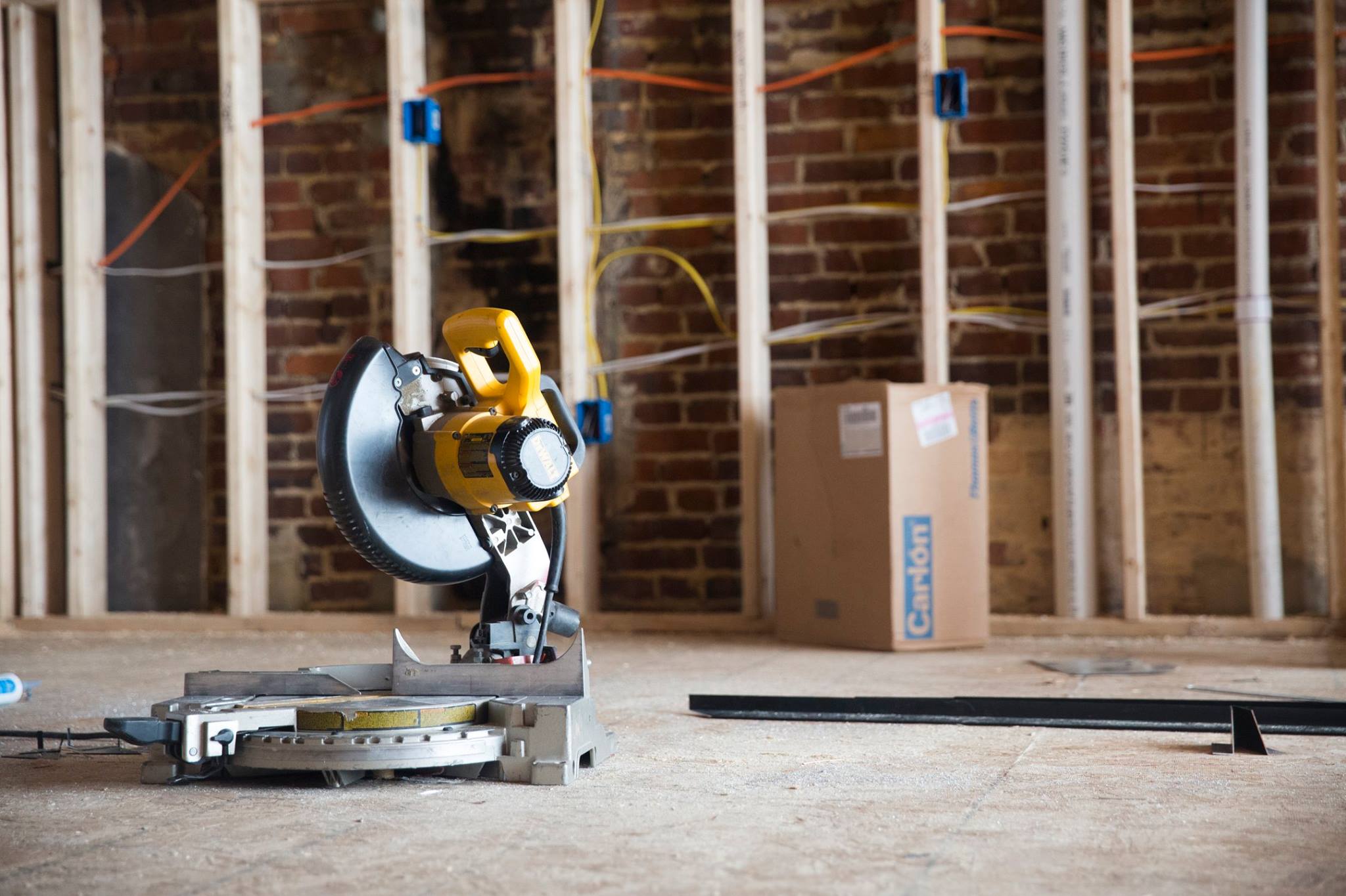 Yellow and black miter saw on floor in an unfinished room with exposed brick wall and wooden studs.