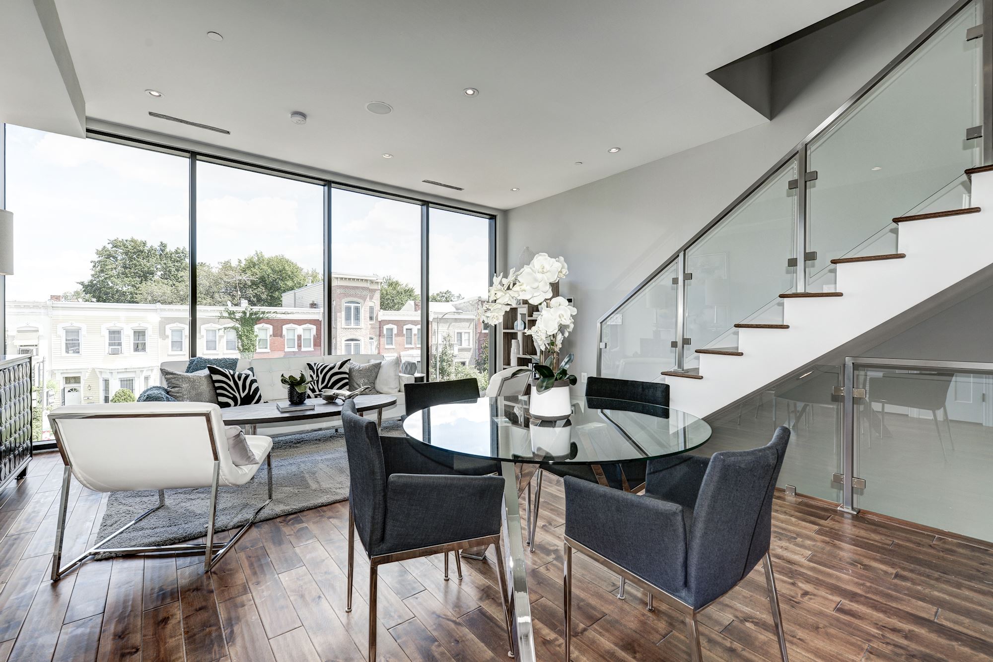 Bright modern living room with large floor-to-ceiling windows, glass dining table with black chairs, a white chair, and a staircase with glass railing.