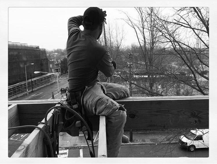 A construction worker wearing a cap and ripped pants sits on a wooden beam overlooking an urban street with bare trees and parked cars.