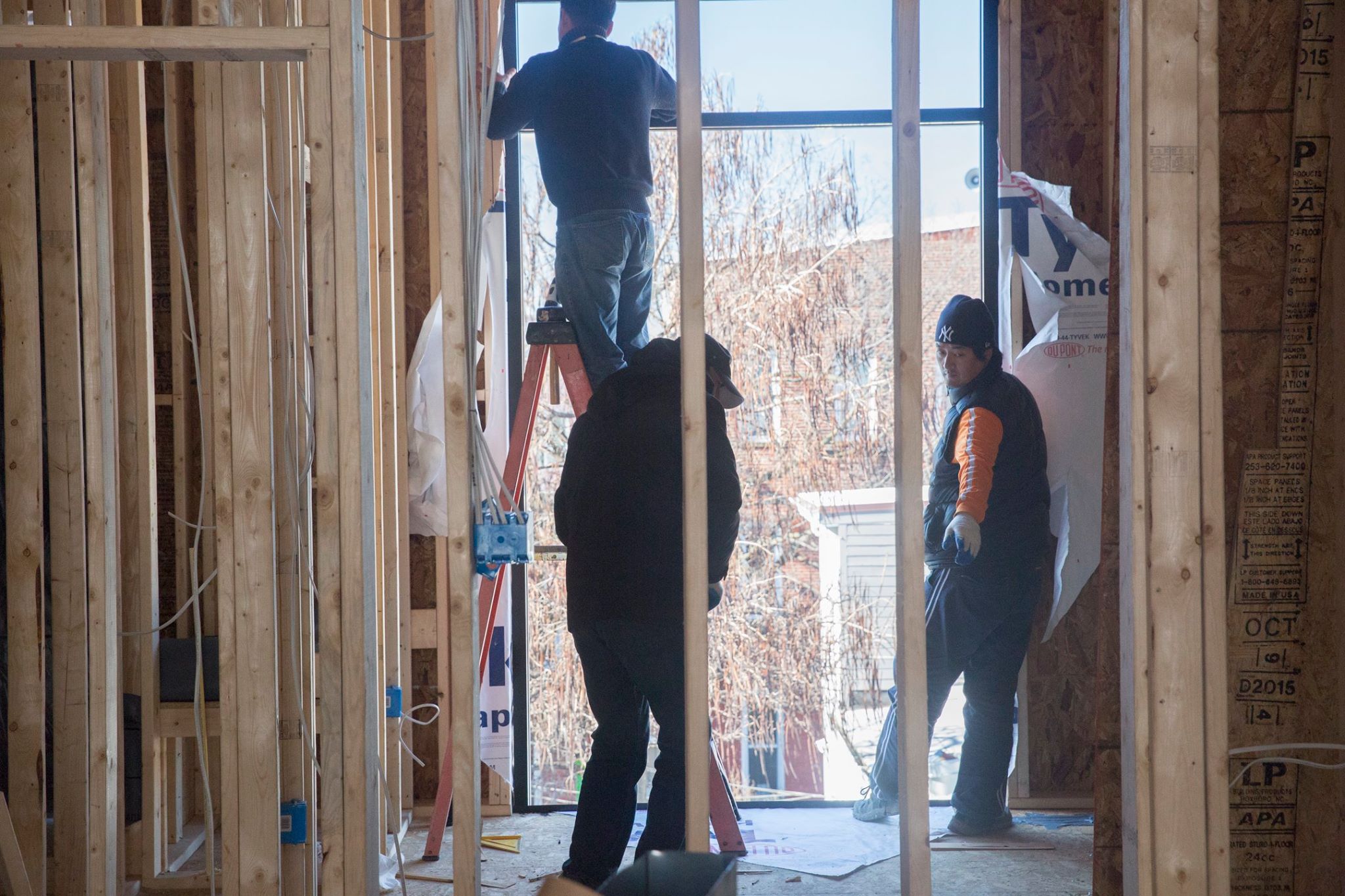 Three construction workers inside a wooden frame structure, one standing on a ladder near a large window opening.