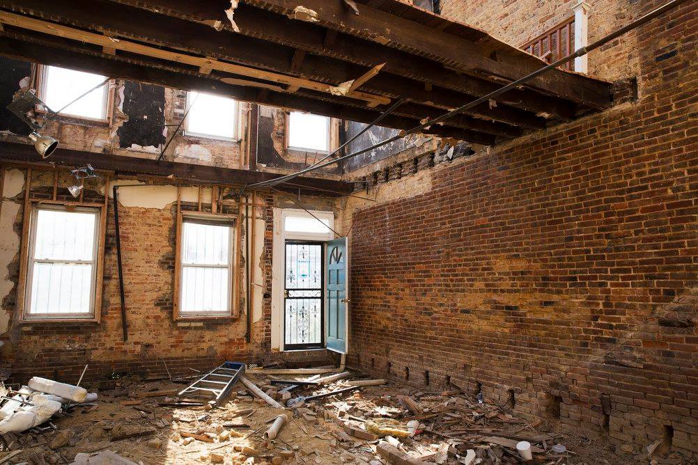 Interior of a dilapidated brick room with exposed beams, broken windows, a partially open door, and debris covering the floor.