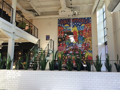 Modern indoor space with white tiled wall, snake plants, a staircase, and a colorful mural with the word 'UNSTOPPABLE'.