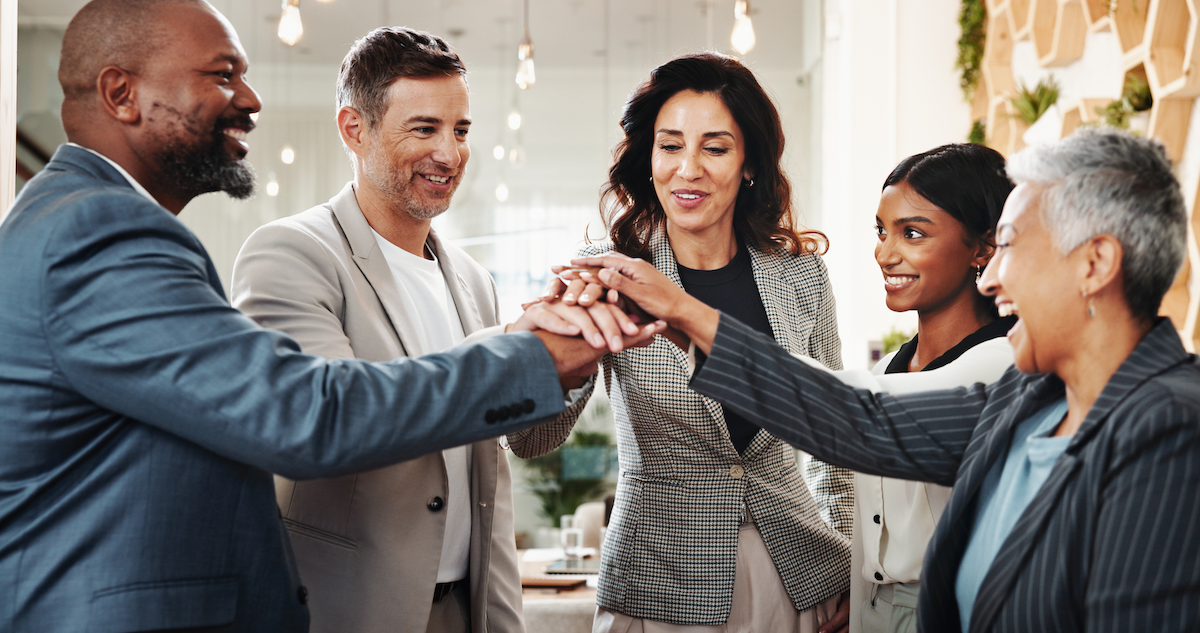 Diverse group of five businesspeople smiling and stacking their hands in a teamwork gesture in an office.