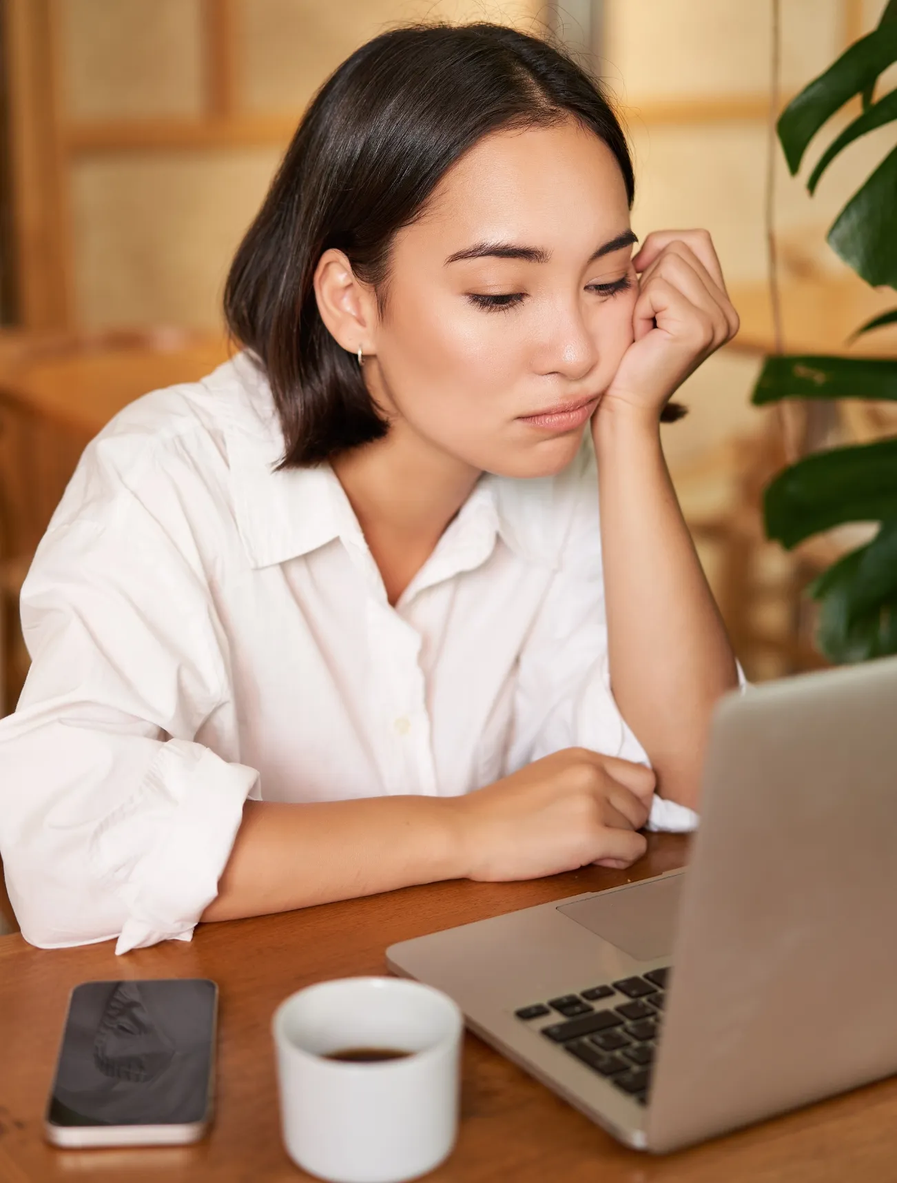 Tired young woman resting head on hand while looking at laptop screen at a wooden table with a coffee cup and smartphone.